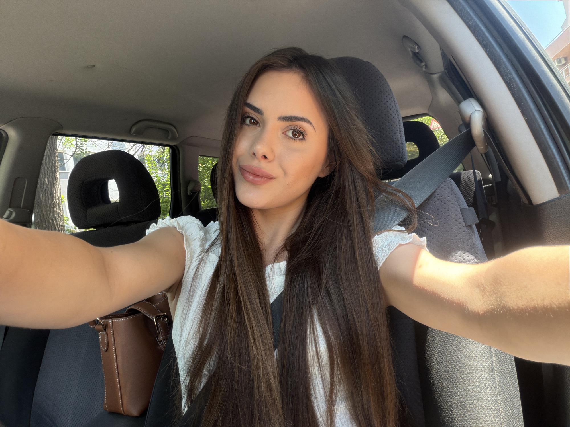 A young woman is depicted taking a selfie inside a car, looking directly at the camera with a soft smile. Her long, straight, dark brown hair is parted in the middle and cascades over her shoulders and chest, largely covering her white top which has ruffled sleeves. She wears makeup, including defined eyebrows, eyeliner, and light pink lipstick.
She is seated in what appears to be the front passenger seat, wearing a dark gray seatbelt across her chest. A small, structured brown leather handbag with light brown stitching and a buckle detail is resting on the car seat next to her.
The car's interior features dark gray fabric seats and headrests, the latter having a distinctive circular cutout. The ceiling and door pillars are light gray. Through the car windows, lush green trees and parts of a building are visible, suggesting an outdoor, possibly urban or suburban, setting. Bright natural sunlight illuminates the scene, creating highlights on her skin and the car's interior. The overall atmosphere is relaxed and pleasant, captured during the daytime. The location context indicates this is in Burgas, Bulgaria. No readable text is visible in the image.