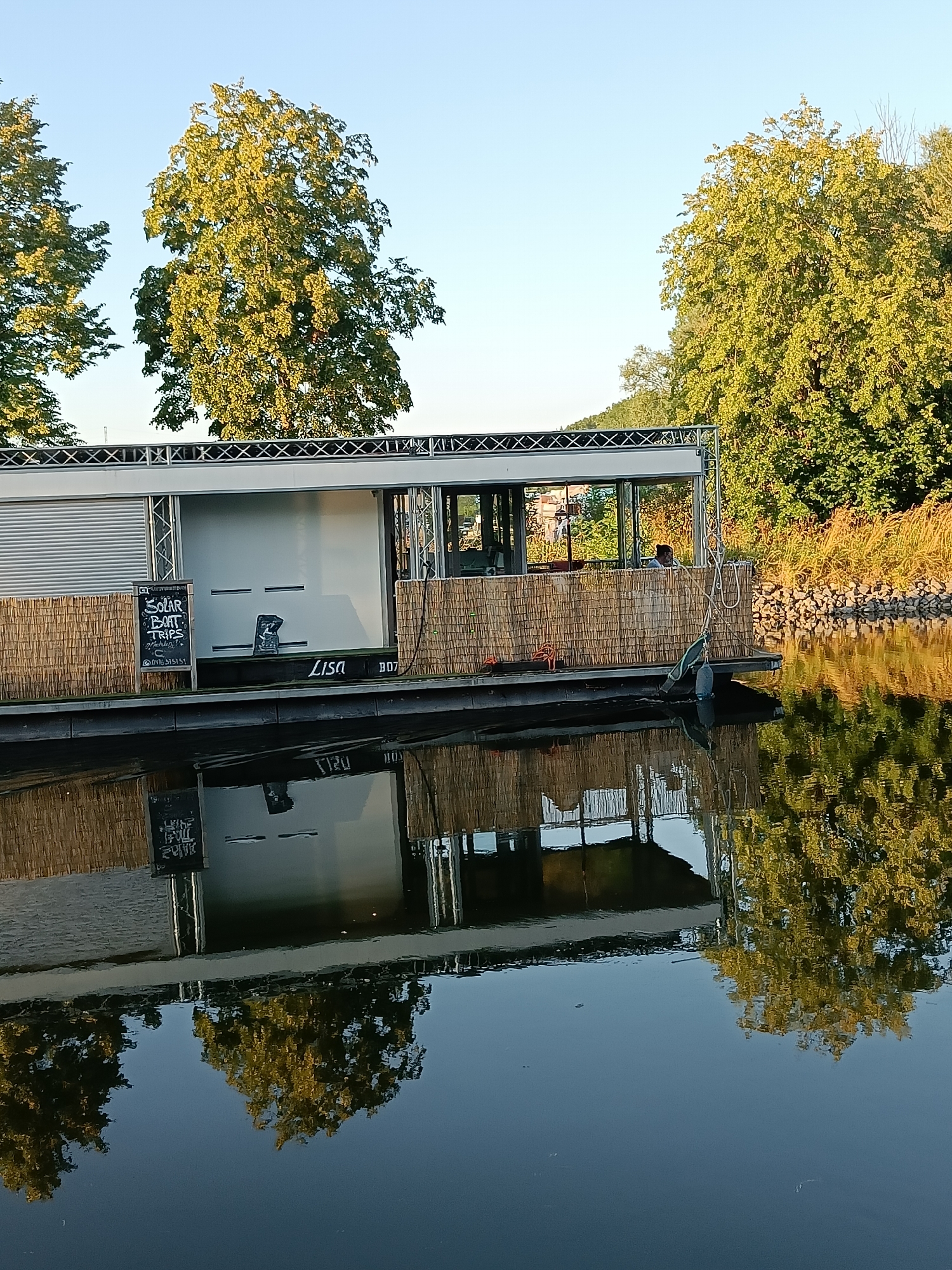 The image depicts a houseboat named "Lisa" docked on a body of water. The houseboat has a modern, rectangular design with large windows and a slatted siding. A sign on the side of the houseboat advertises "Solar Boat Trips." A person is visible through one of the windows, seated inside. The houseboat is surrounded by trees and vegetation, with a clear sky in the background. The reflection of the houseboat and trees is visible in the calm water. The scene appears to be set in a park or natural area, possibly along a canal or river. The city of Haacht, Belgium, is known for its green spaces and waterways, making it a plausible location for such a scene.