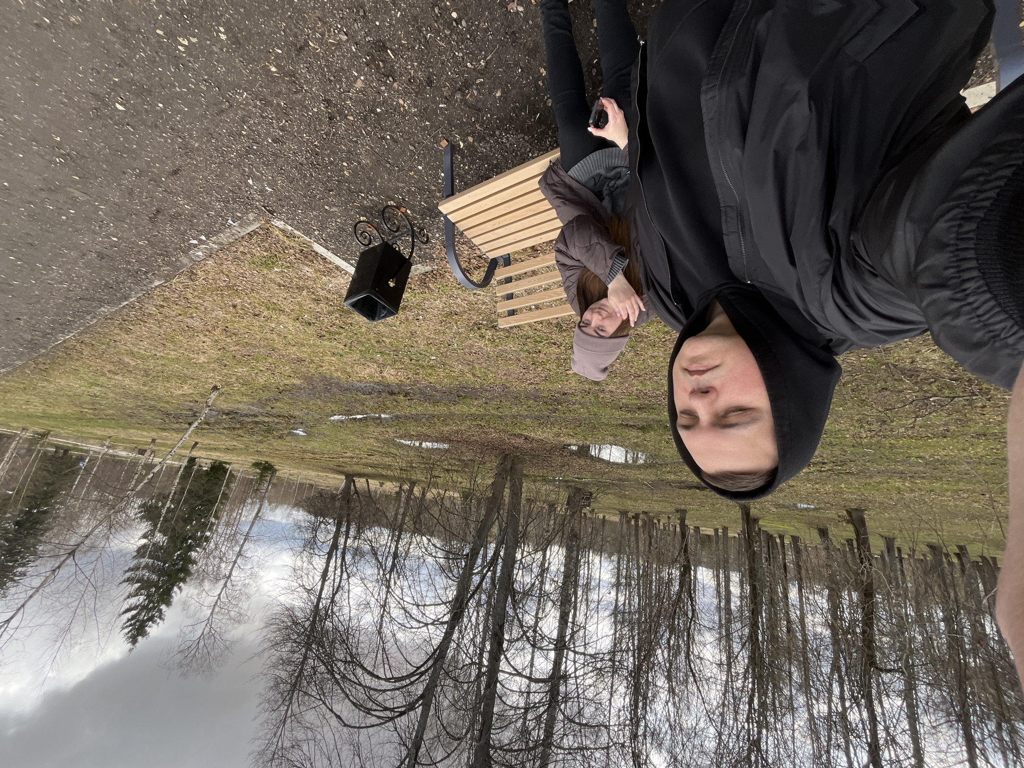 Two individuals are situated in a park-like environment in what appears to be the city of Vysokovsk, Russia. One person, wearing a black hooded jacket, is positioned in the foreground, seemingly holding the camera for a selfie. The other individual is seated on a wooden bench, wearing a brown jacket and a purple-grey beanie. The bench is located on a grassy area with scattered fallen leaves. A black trash receptacle is placed next to the bench. The background consists of a line of bare trees and a cloudy sky, suggesting it could be early spring or late autumn. A paved walkway is visible to the left.