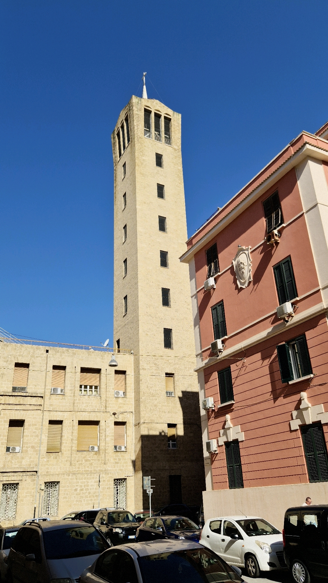 A low-angle, slightly tilted shot captures the clear blue sky above a cluster of buildings in Taranto, Italy. Dominating the left side of the frame is a tall, slender, light-colored stone tower with a pointed steeple. The tower features several evenly spaced rectangular windows and a section near the top with arched openings. To the right, a multi-story building with a salmon-pink facade and white trim is angled towards the viewer. This building has dark-shuttered windows, air conditioning units mounted below some of them, and decorative architectural elements above the upper-story windows. In front of these buildings, a street level view shows several parked cars, including a silver sedan, a black SUV, a dark blue sedan, and a white compact car. The lower portion of a tan stone building is visible on the far left, with numerous windows, some fitted with blinds, and air conditioning units. A street sign is visible near the center of the frame. The overall impression is an urban street scene bathed in bright sunlight, casting distinct shadows.