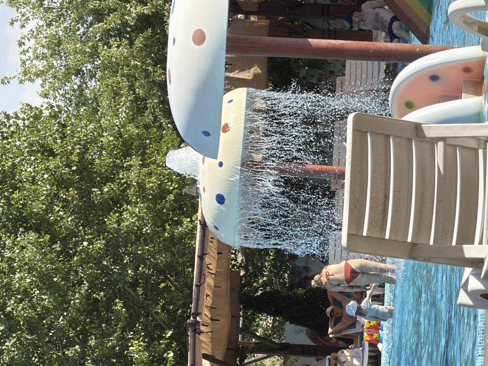 The image depicts an outdoor water park in Sociteni, Moldova, on a sunny day. In the foreground, a white plastic lounge chair is partially visible, facing away from the camera. The middle ground features a swimming pool with clear blue water. A woman in a red bikini stands in the water, with her back to the camera, and a young child wearing a white sun hat and a light blue swimsuit is in the pool near her.
To the left of the woman, a man is seated in a lounge chair under a thatched canopy, wearing a baseball cap. Behind the lounge chair, a green evergreen tree stands.
In the center and right of the image are two mushroom-shaped water features. The one on the left is yellow with red and blue polka dots and is actively spraying water downwards. The one on the right is white with red and blue polka dots. Behind these water features, more lounge chairs and a wooden structure are visible. Further to the right, a man wearing a dark baseball cap and dark swim trunks is seated by a colorful slide, looking towards the left. Lush green trees with dense foliage form the backdrop of the scene, and the sky is a bright blue with some visible clouds.