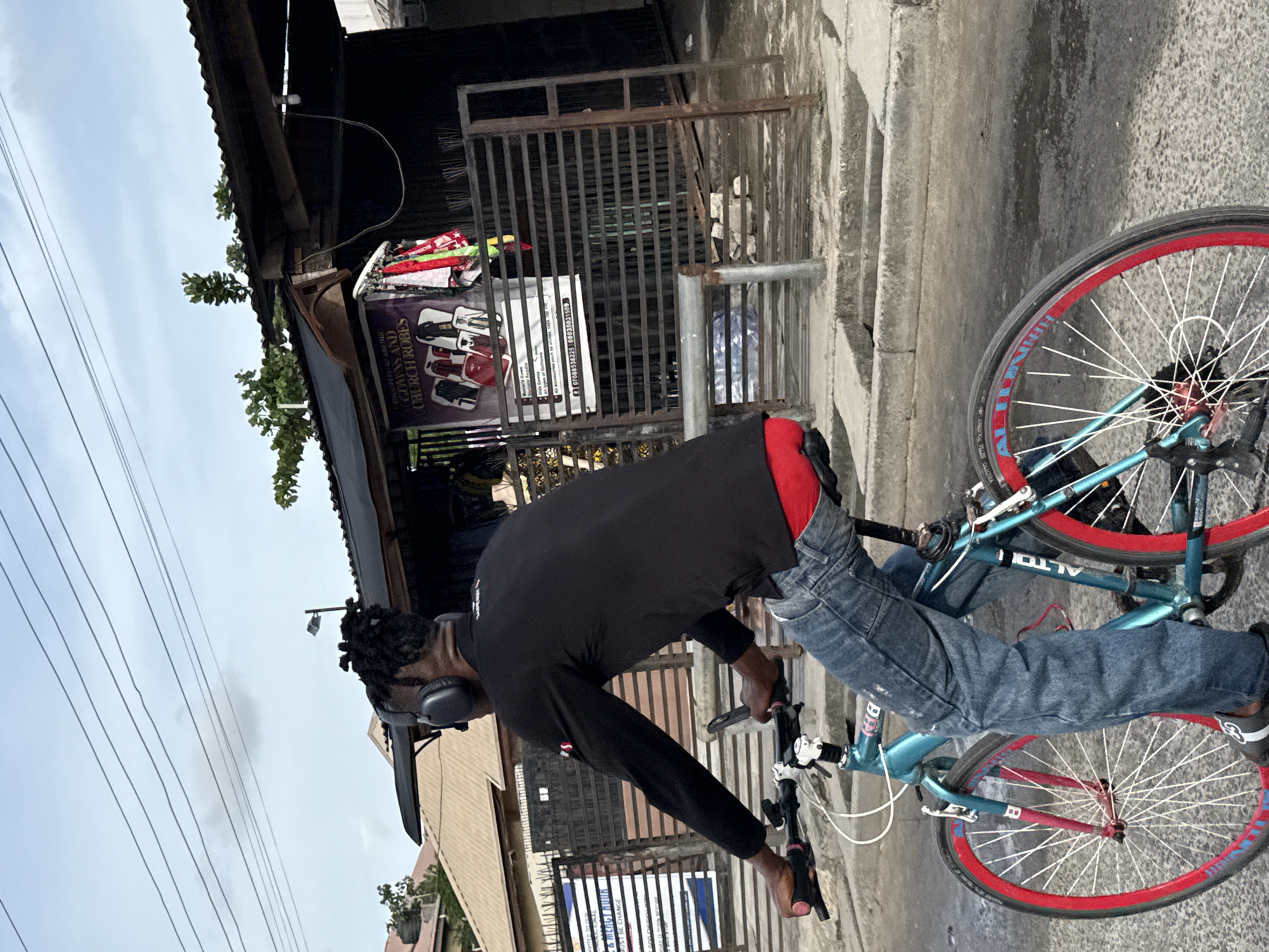 This vertical image captures a person on a bicycle in what appears to be an urban street in Ikorodu, Nigeria. The central figure, viewed from behind, wears a black long-sleeved shirt, blue jeans, and has red underwear visible above their waistline. They sport dreadlocks and are wearing black over-ear headphones. The person is either riding or standing astride a light blue bicycle with distinctive red rims and white spokes. The bicycle frame features "ALTO" and "BH" branding.
The background reveals a bustling street environment. To the right, there are various buildings with corrugated metal roofs and concrete walls, some featuring metal security gates. Overhead, multiple power lines crisscross an overcast sky. A prominent purple and white sign is visible on a building, advertising "conventians and seminar" for "GOWNS AND CHURCH ROBES," along with "All accessories for church programs, cap & tassel." A phone number, "07066583225 08023503159," is also listed. Another, partially obscured white sign behind the gates appears to mention "TAILORING & MENU..."
The time of day is daytime, indicated by the overall brightness, though the sky is cloudy, suggesting an overcast condition rather than direct sun.