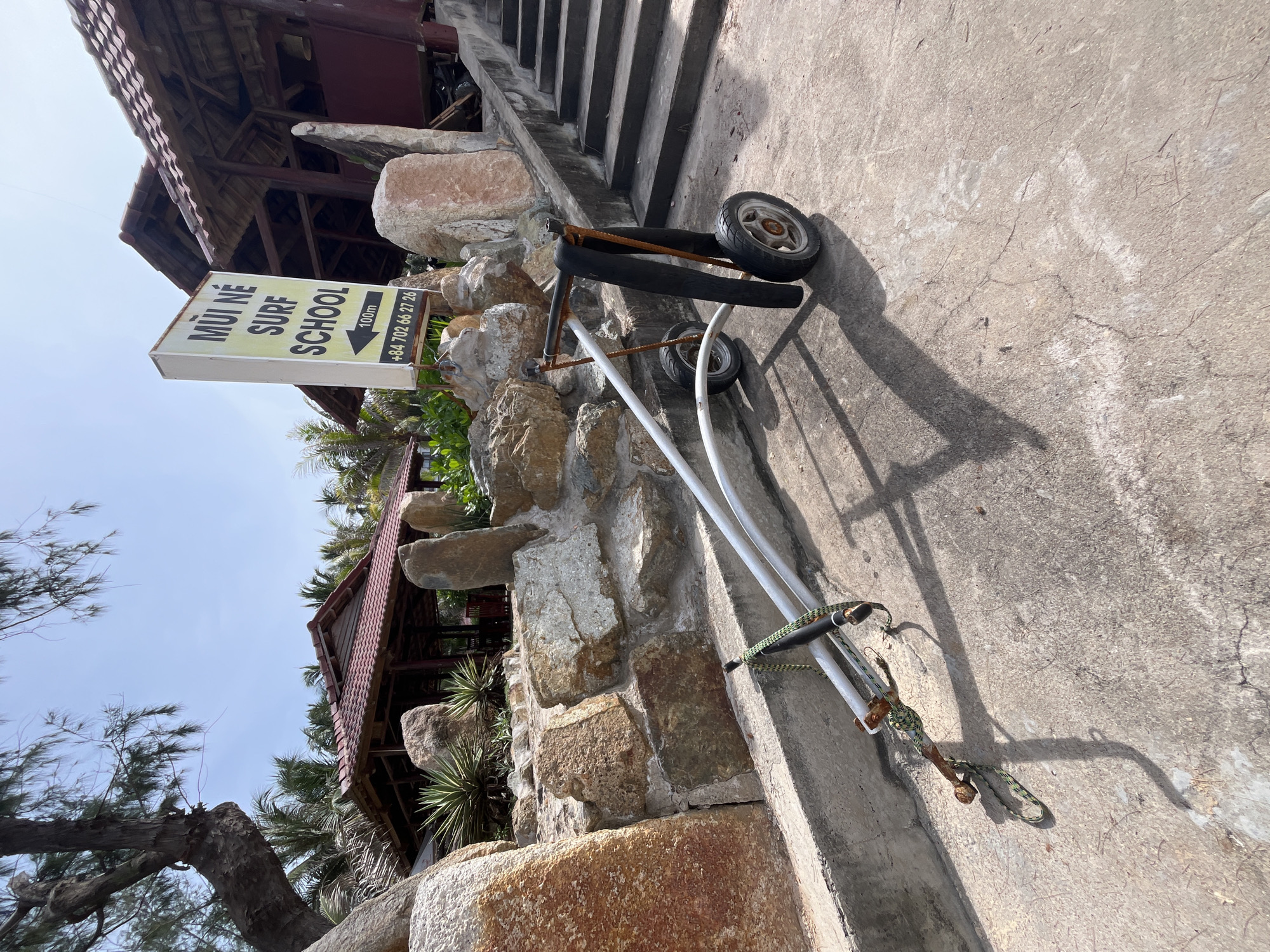 The image depicts an outdoor scene in Phu Thuy Ward, Vietnam, likely at a surf school. In the foreground, a weathered trolley with two wheels stands on a concrete surface. The trolley appears to be constructed of metal tubing, with a black material draped over its handles. A rope is tied to one of the trolley's supports. Behind the trolley, a stone wall is visible, constructed from large, irregularly shaped rocks. Beyond the wall, a wooden building with a red tile roof is partially visible. A sign with yellow lettering on a white background is mounted on a pole, reading "MUI NE SURF SCHOOL" with an arrow pointing left and the text "100m" and a phone number. Several palm trees and other vegetation surround the structures. The sky is clear and blue. To the right of the building, a concrete staircase ascends. The overall impression is a casual, slightly rustic environment associated with a beachside activity.