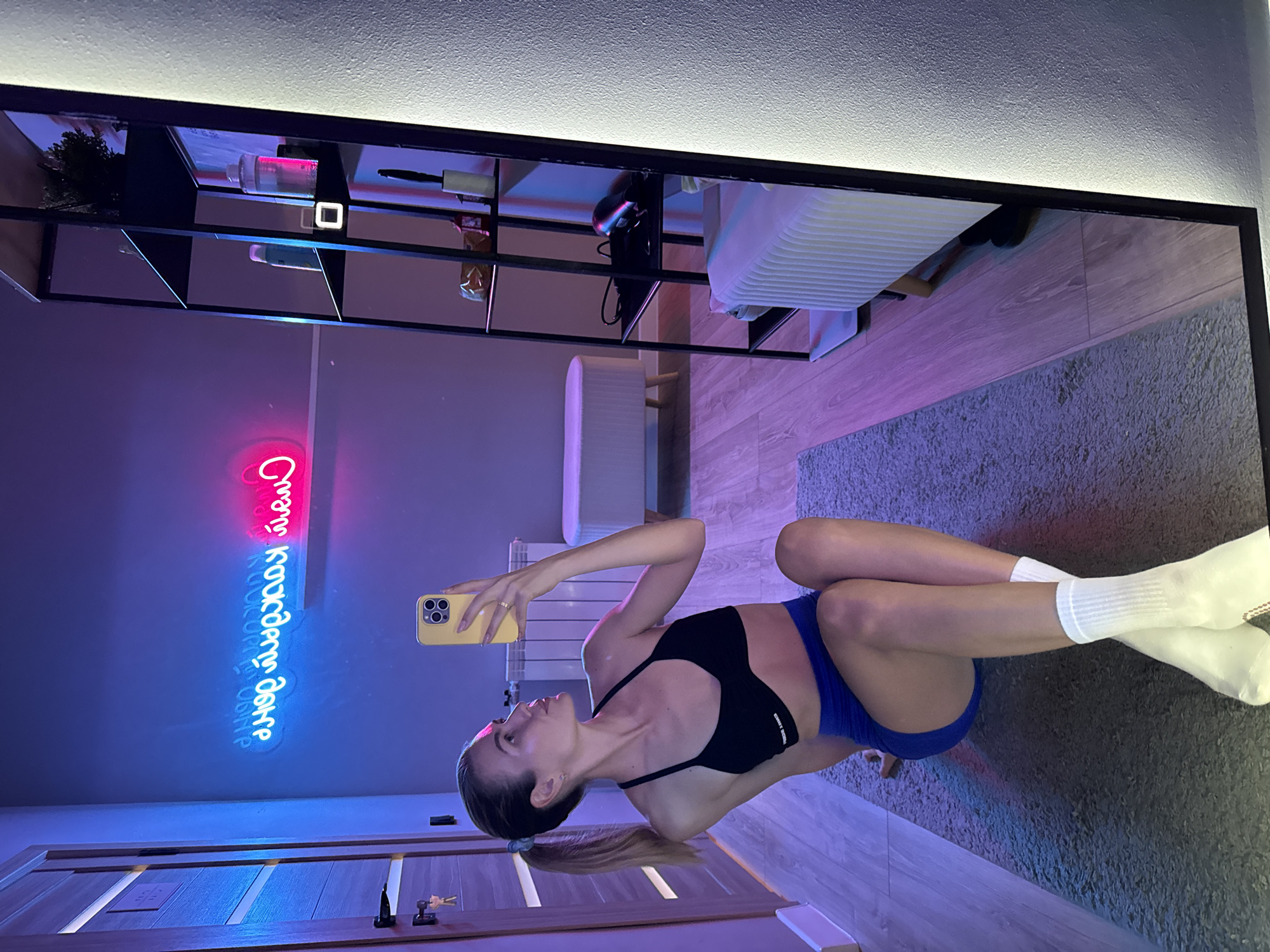 A woman is posing for a mirror selfie in an interior setting. The room features a door in the lower left, a grey carpet on the floor, and a neon sign displaying what appears to be a message in a non-English alphabet. Above, there is a wall-mounted shelf with various items, including bottles and what looks like gaming headphones. The woman is wearing a black sports bra and blue shorts, complemented by white socks. She holds a yellow smartphone up towards the mirror, with the reflection showing the full image. The room is lit with a blue and pink hue, adding to the overall ambiance. The location is Unknown, Unknown.