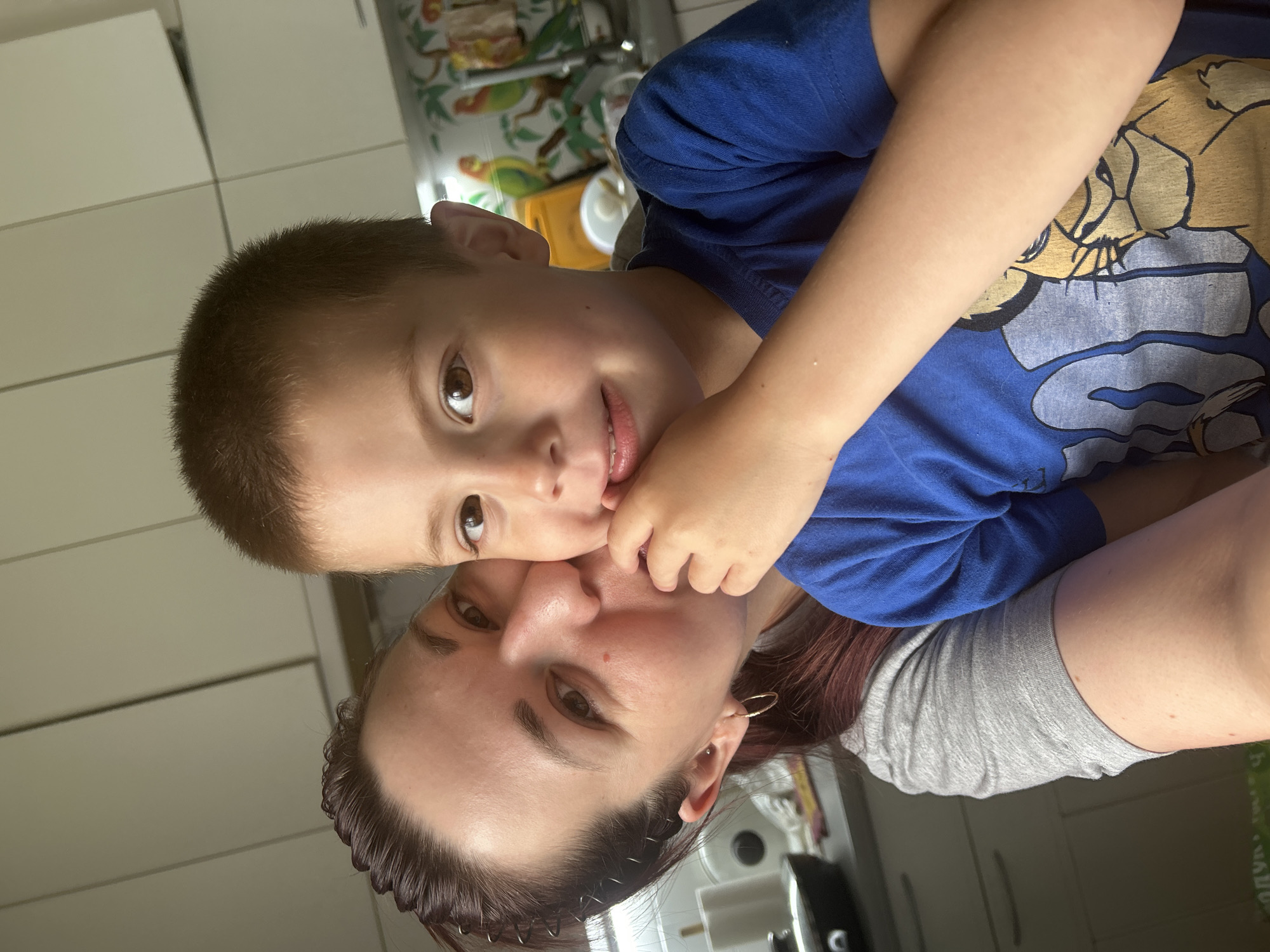 This media file is a close-up, vertical, eye-level shot of a woman and a young boy. The woman is in the foreground, with the boy embracing her from behind, leaning into her face. Both subjects are smiling. The woman has dark hair pulled back, and is wearing a grey t-shirt and hoop earrings. The boy has short, dark hair and is wearing a blue t-shirt with a graphic of characters from Disney's The Lion King. The background appears to be a kitchen, with white cabinets and a colorful wallpaper featuring birds. The lighting suggests it is daytime. The overall mood is affectionate and joyful. There is no visible text in the image.