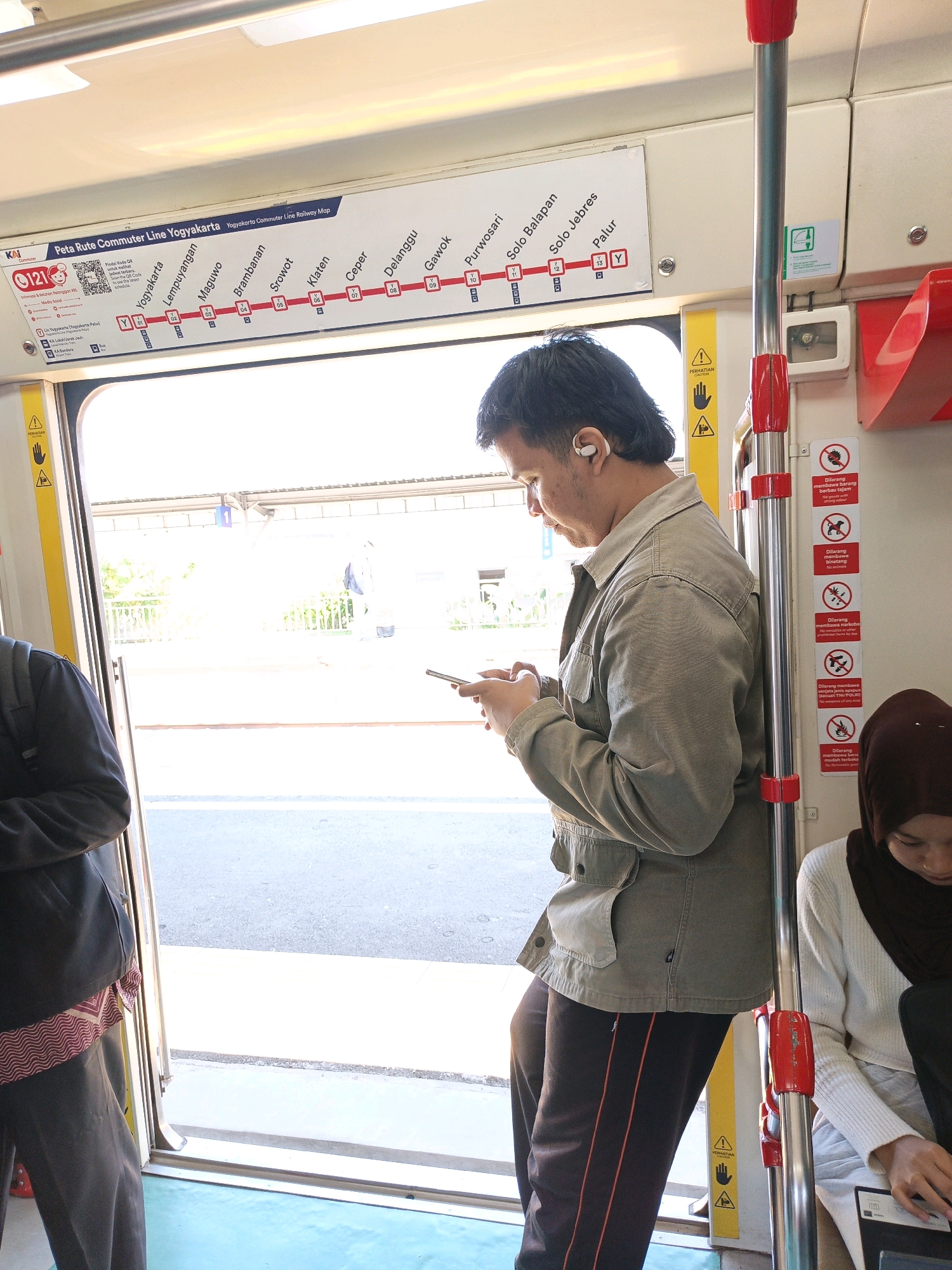 The image depicts the interior of a train carriage, possibly a commuter train, with a focus on the doorway and the passengers present. A man wearing a grey jacket and dark pants with an orange stripe is standing near the doorway, looking down at his mobile phone and wearing wireless earbuds. To his left, another person in a dark jacket and patterned scarf is partially visible, facing away from the camera. To the right, a woman wearing a white sweater and a dark brown hijab is seated and looking down at something in her lap.
The train carriage interior features a map of the "Yogyakarta Commuter Line Railway Map" displayed above the doorway, listing various stations. Prominently displayed on the wall next to the man are several red and white prohibition signs, indicating items or actions that are not allowed on the train, such as animals and certain types of goods. A yellow warning sign with hand icons is also visible.
The view through the open doorway shows a bright, sunny outdoor environment with a train platform and greenery in the distance. The exterior appears to be a station, possibly in or around Klepu, Indonesia, given the mention of the Yogyakarta Commuter Line. The floor of the carriage is a light blue color.