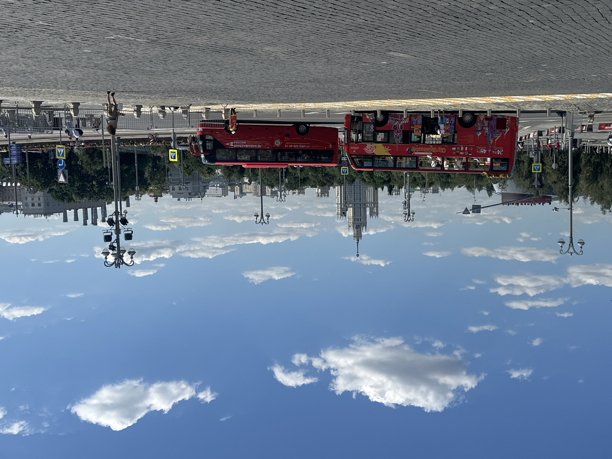 In an urban setting under a clear blue sky with scattered white clouds, two red double-decker buses are parked on a cobblestone street. The bus on the left, numbered 24 and 48, displays "City Sightseeing" and colorful depictions of buildings. The bus on the right has "Незабываемая МОСКВА" written on its side, indicating it is a Moscow sightseeing bus. Several people are visible, including passengers on the upper deck of the left bus, a man standing near the open door of the right bus, and a person in a yellow vest near the right bus. In the background, a tall, ornate building with a spire is prominent, along with other city buildings and trees. Streetlights and traffic signs are also present. The scene appears to be a tourist area in Moscow, Russia.