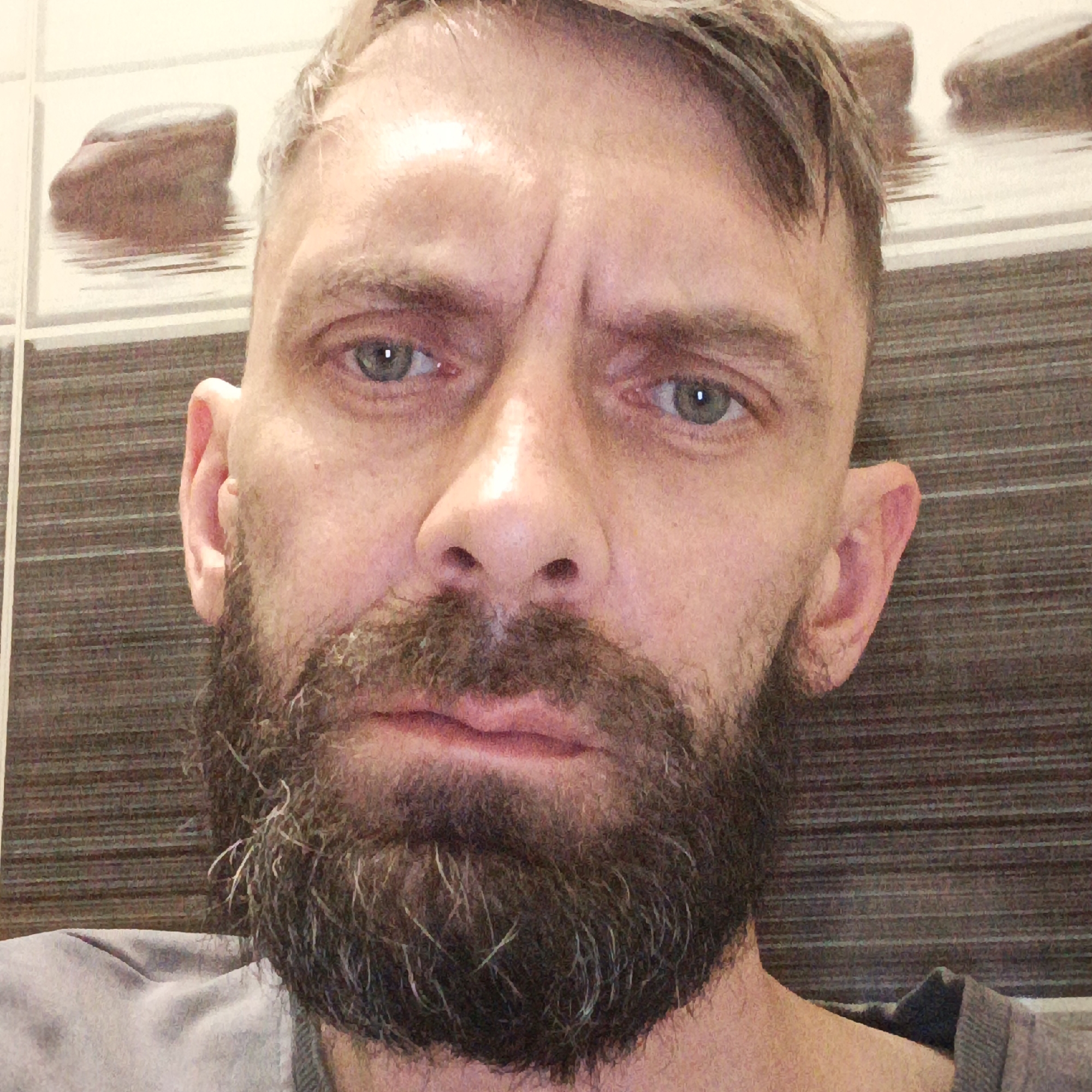 A close-up shot of a middle-aged man with a salt-and-pepper beard and short, faded hair. He has a furrowed brow and appears to be looking directly at the camera. The background consists of tiled walls, with images of rocks or stones embedded within the tile design. The man is wearing a gray shirt. The location information (Марковский, Russia) is not discernible from the image.