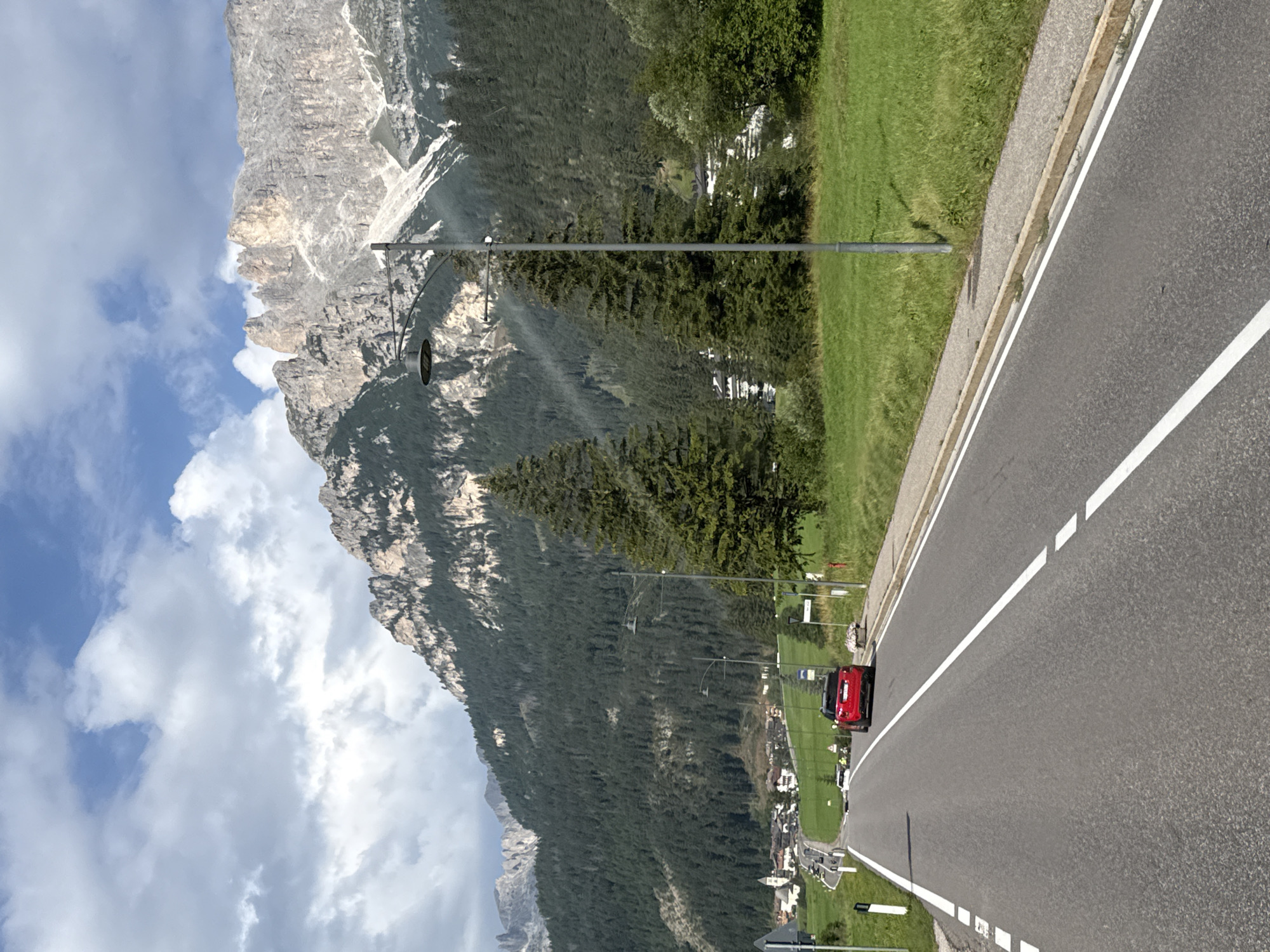The image shows a road leading towards the town of Mazzin in Italy, with a mountainous backdrop. A red car is visible on the road, approaching the viewer. The surrounding environment is characterized by lush green fields and dense pine forests on the slopes of the mountains. The sky is partly cloudy. In the distance, the town of Mazzin appears nestled among the hills, with buildings and a church steeple visible. The road itself is paved and marked with white lines. A lamppost stands on the right side of the road, and there are some trees along the roadside.