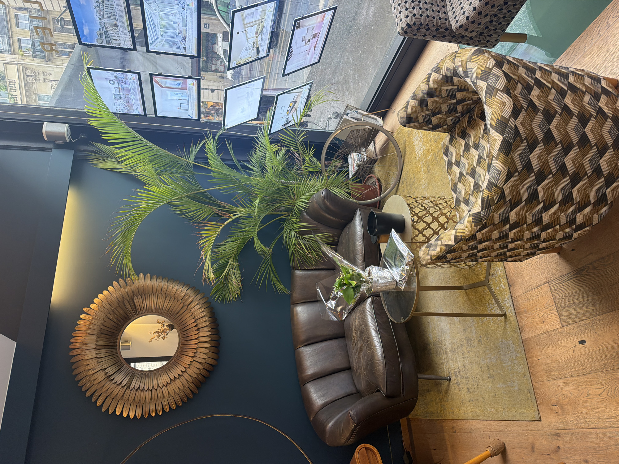 The image depicts an interior scene with an assortment of decor items. There is a brown leather couch, a round decorative gold mirror, a large potted palm plant, and a glass-topped side table with a bouquet of flowers. An armchair with a patterned fabric sits nearby. In the background, there is a window display with multiple real estate advertisements. The scene is indoors, possibly a real estate office or staging area, with hardwood floors. The location is likely in Paris, France, suggested by the architectural style visible in the real estate advertisements displayed in the windows.