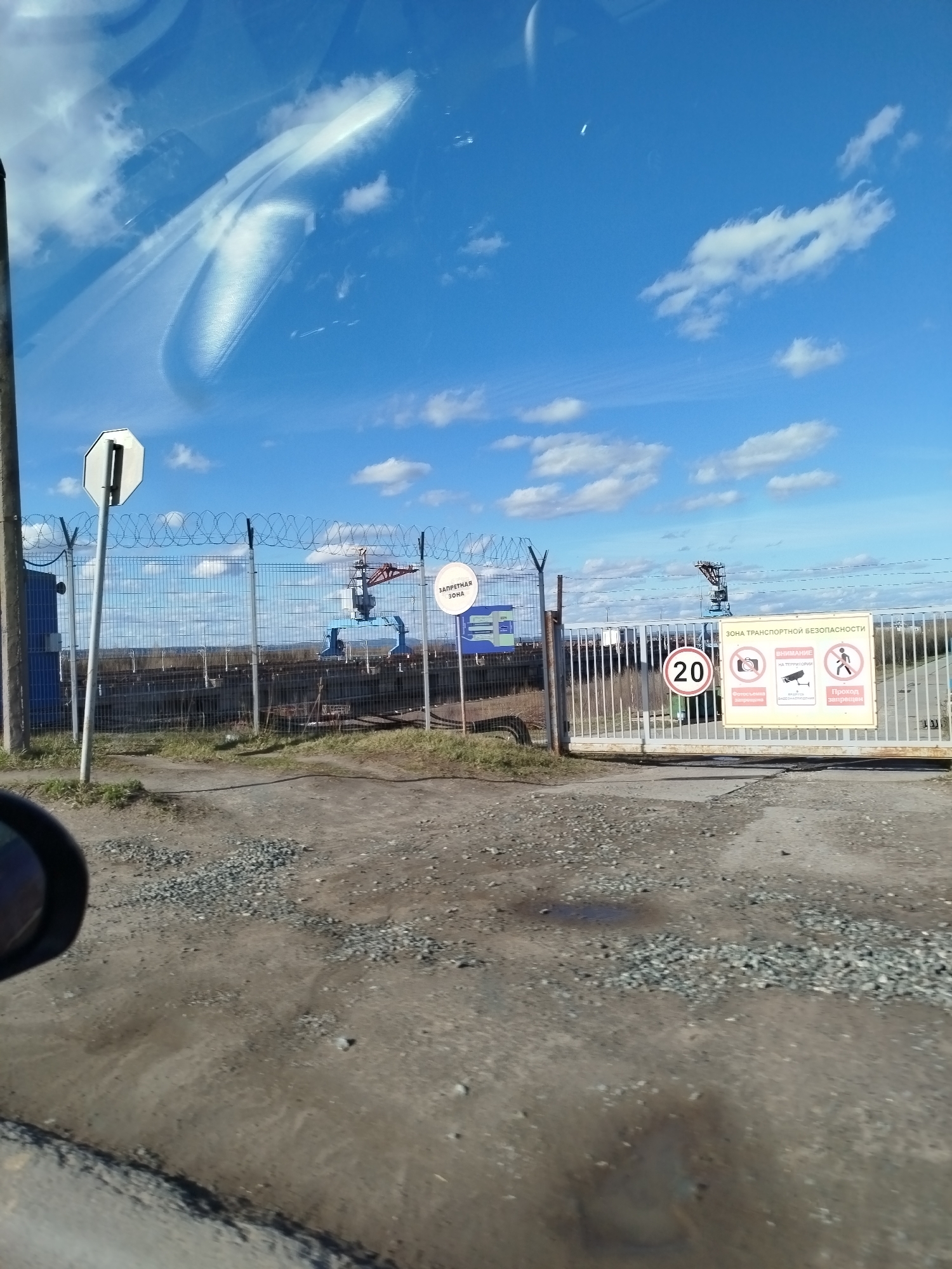 The image shows the entrance to an industrial facility or restricted area. There's a chain-link fence topped with barbed wire, various warning signs in Russian, including a speed limit sign of 20 (likely kilometers per hour). The signs indicate a transportation security zone, forbidding photography and pedestrian access. A large crane is visible inside the fenced area. The foreground shows a dirt road or unpaved area. The sky is blue with scattered clouds. The image appears to have been taken from inside a vehicle, as part of the side mirror and windshield reflections are visible.
Based on reverse image searches and the presence of the signs in Russian, it is possible that this could be in Chaikovsky, Russia. Specifically, the "Chaikovsky-Textile" industrial complex.