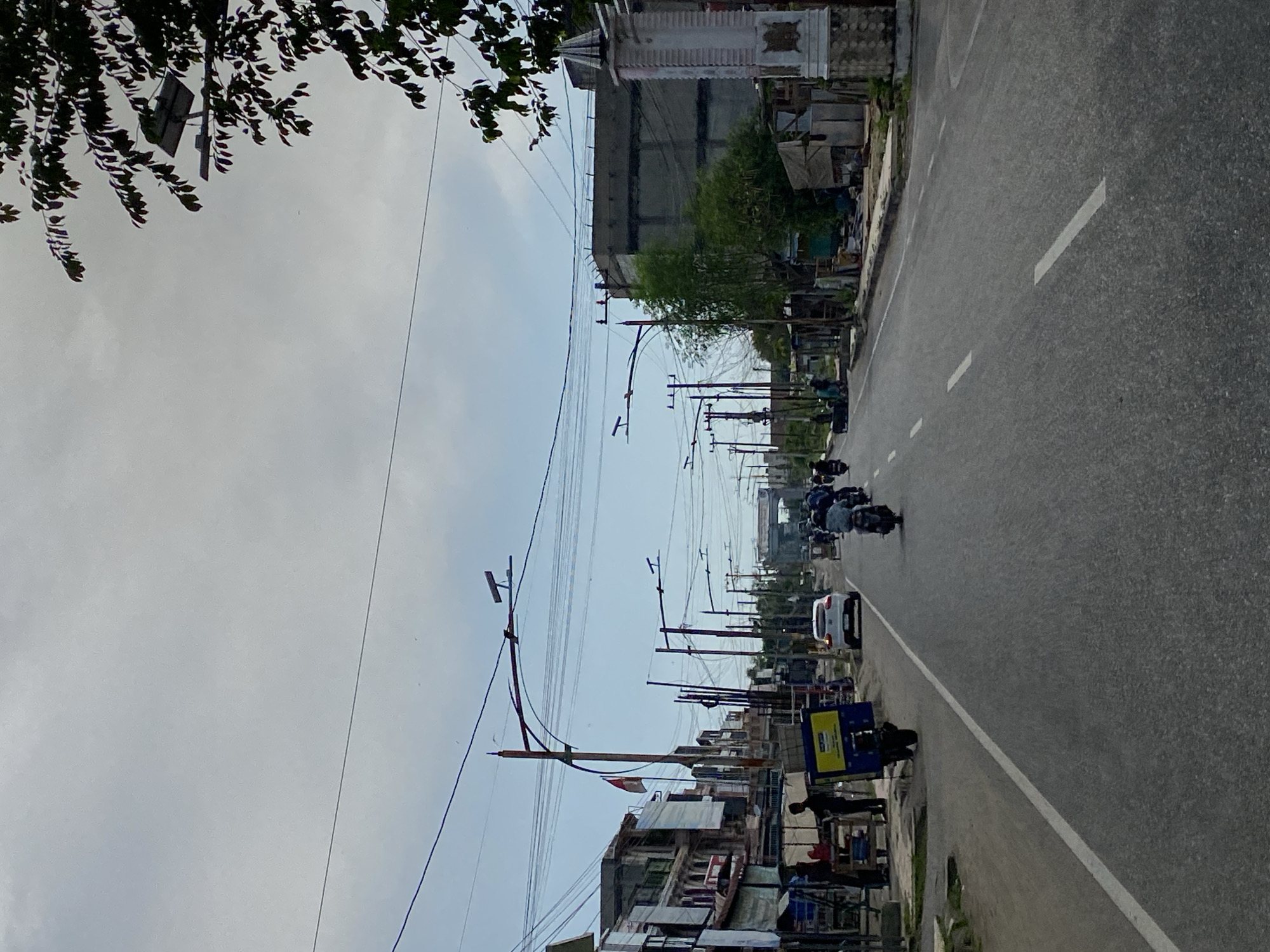The image captures a street scene in Lhokseumawe, Indonesia, on a cloudy day. The road is paved and marked with white lines, indicating a moderately busy thoroughfare. Numerous utility poles with a complex network of wires crisscross the sky, common in urban Indonesian settings. On the left side of the road, there are low-rise buildings with shops and stalls, some displaying signage. A few individuals are visible near these establishments, some appearing to be vendors or shoppers. A white car is traveling down the road, and several motorcycles are also present, with some riders wearing helmets. On the right side, there are more buildings, including a larger, more industrial-looking structure and smaller, temporary shelters. A decorative pillar or monument stands near the right edge of the frame. The presence of the Indonesian flag on a pole suggests a national or public holiday, or simply a display of patriotism. The overall impression is of a typical urban street in a coastal city in Indonesia, likely experiencing normal daily activity.
