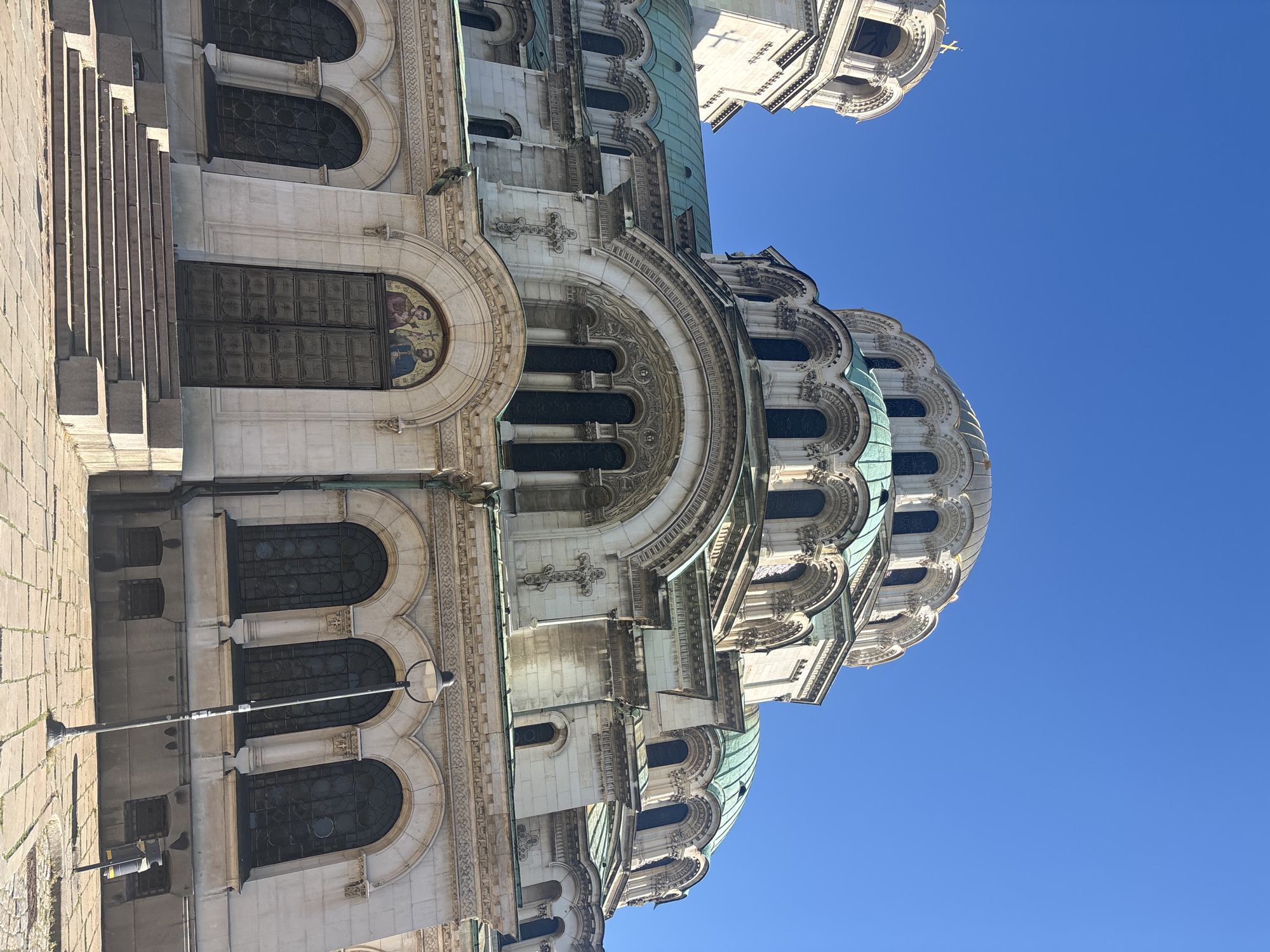 The image features the Alexander Nevsky Cathedral in Sofia, Bulgaria, viewed from a low angle. The cathedral is a grand structure with ornate detailing, domes with a greenish tint, and arched windows. The clear blue sky is visible in the background, contrasting with the light-colored stone of the cathedral. The image primarily focuses on the cathedral's exterior architecture, with no people present in the frame.