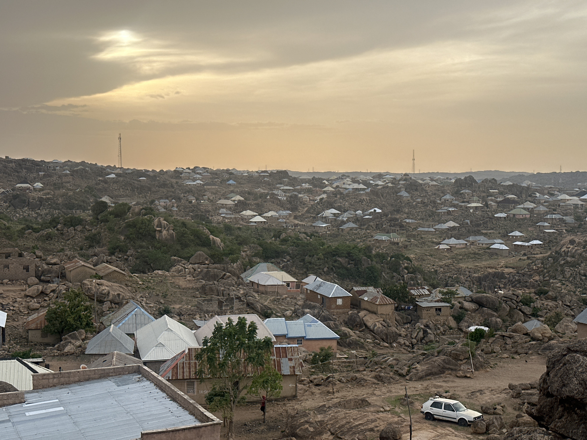The scene is a high-angle view of a section of Jos, Nigeria. The landscape is characterized by numerous large, gray rocks interspersed with small buildings and houses. The buildings have metal roofs of various colors, including silver, blue, and pink. Scattered vegetation is present, including trees and shrubs.
In the foreground, there are more buildings with corrugated metal roofs. A few trees are visible in front of these buildings, and a person is standing near one of the trees. A white car is parked on the rocky ground.
In the background, the scene extends into the distance, with more houses and rocks stretching to the horizon. Several tall communication towers are visible in the distance against a pale yellow and gray sky. The overall environment suggests a residential area built within a rocky landscape.