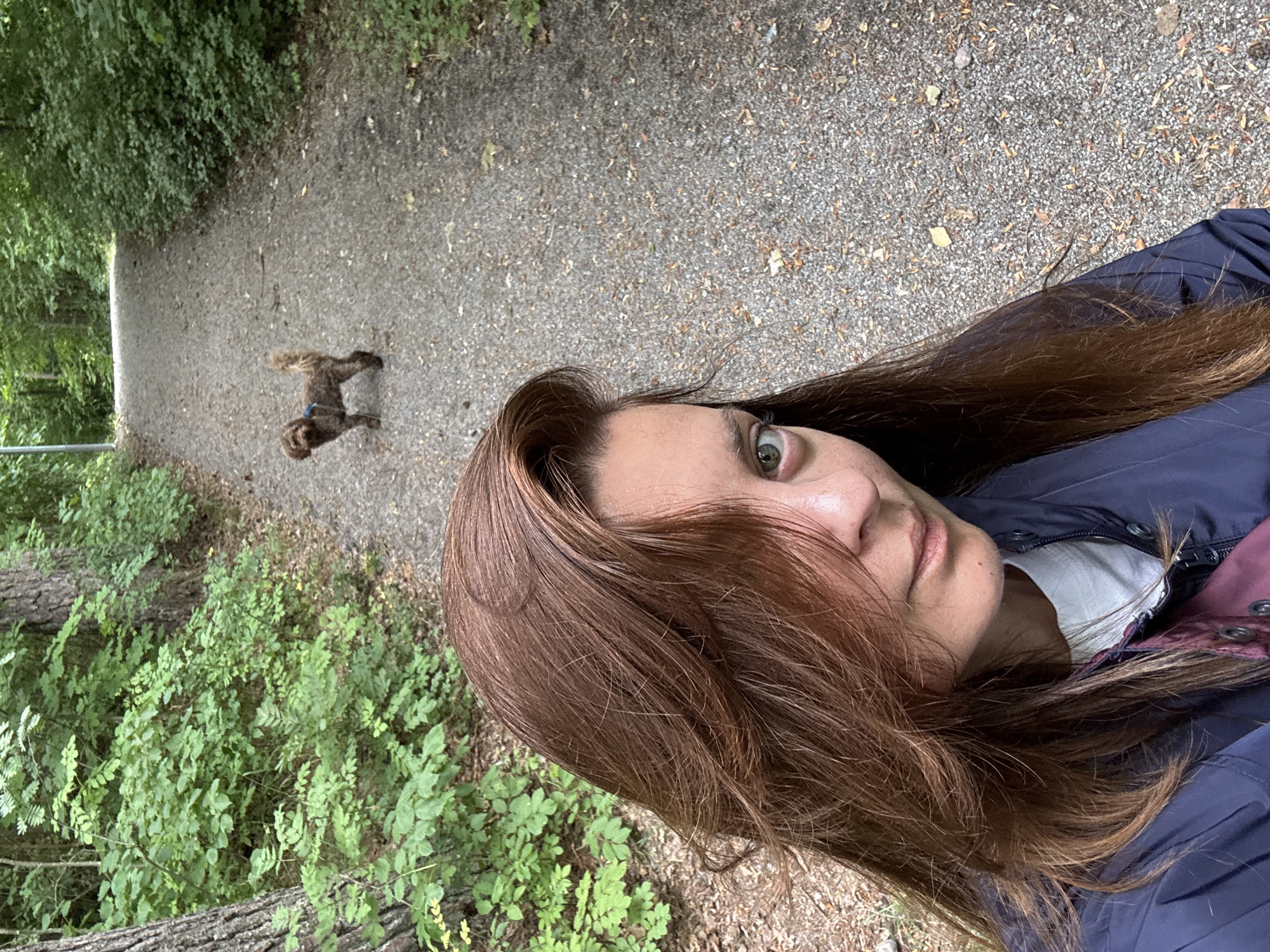A woman with long, auburn hair is taking a selfie on a gravel path in a wooded area. The woman is wearing a dark blue jacket over a white t-shirt. Her eyes are green, and she is smiling slightly. In the background, a small, brown dog with a curly tail stands on the path, facing away from the camera. The path is surrounded by lush green trees and bushes. The image appears to have been taken in the city of Åkersberga, Sweden, based on the context provided.