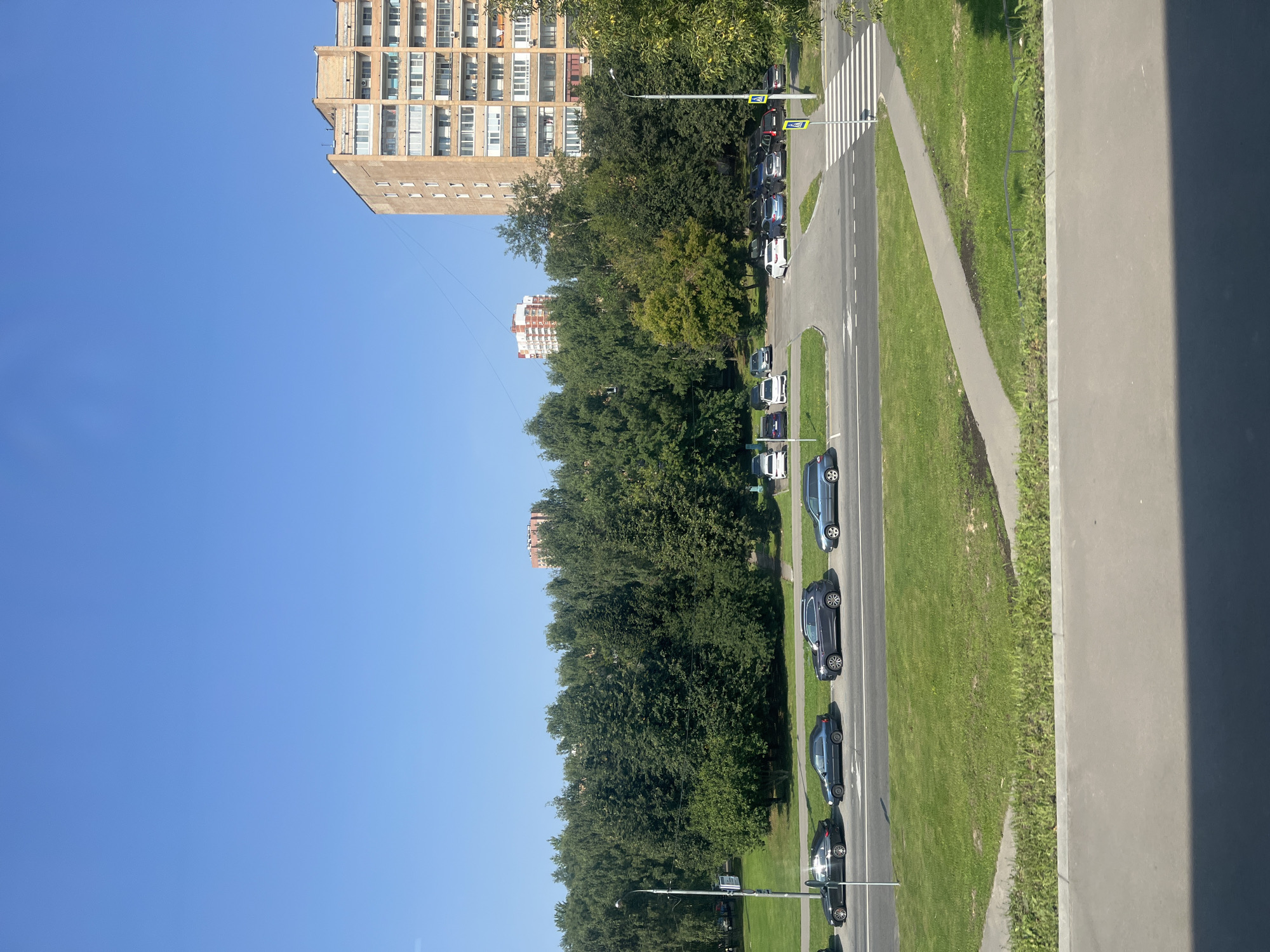 The image captures a sunny day scene in Moscow, Russia, featuring a typical urban landscape. In the foreground, a paved walkway runs parallel to a road where several cars are parked. Beyond the road, a vast expanse of green lawn is visible, dotted with trees and a pedestrian crossing. Further into the background, a large, multi-story apartment building stands tall, its facade a light brown color, indicating residential architecture common in Moscow. Adjacent to it, another apartment building with a more modern design, featuring red and white elements, is visible. The sky is clear and blue, suggesting pleasant weather. The overall impression is one of a residential area in a Russian city, with a focus on apartment living and green spaces.