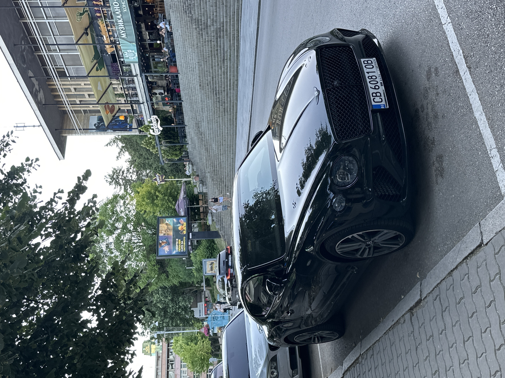 A black Bentley Continental GT is parked on the side of a street in Veliko Tarnovo, Bulgaria, directly in front of a building with outdoor seating for multiple establishments. The car is facing the viewer, with its front grille and headlights prominently displayed. The license plate reads "CB 6081 OB". In the background, a large outdoor staircase leads up to the building, which has multiple levels of outdoor seating areas. Several people are visible seated at tables in these areas, and others are walking up the stairs. There are trees lining the street and the background. A billboard advertising something with Bulgarian text is visible on the left side of the image. The scene appears to be a sunny day in an urban setting.