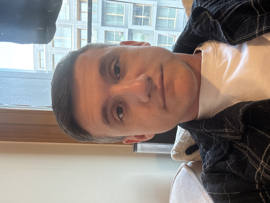 A male individual, approximately 25-35 years of age, with short, dark hair and a neutral expression, is depicted in a medium close-up, looking directly at the camera. He is wearing a white crew-neck t-shirt beneath an open dark-colored, possibly black or dark blue, plaid shirt with light-colored stitching forming the pattern. He is seated indoors, with the upper portion of a light-colored sofa or armchair cushion visible on the left and a darker, textured cushion on the right, indicating he is reclining or seated comfortably.
To the left, a plain light-colored wall is visible, along with a portion of a wooden window frame. Behind his right shoulder (viewer's left), a dark-colored, vertical appliance with a transparent reservoir is partially visible. The background consists of a large window offering a view of a dense urban environment. Outside, multiple multi-story residential buildings of contemporary design are visible under an overcast sky. These buildings feature grey facades, large rectangular windows, and visible balconies or loggias. Their uniform, modern architectural style and close proximity indicate a high-density urban area.
Based on the modern high-density residential architecture characterized by grey facades and repeated window/balcony patterns, the location is consistent with areas of significant recent urban development in **Warsaw, Poland**, particularly within districts like **Wola**, known for its contemporary apartment complexes and urban renewal.