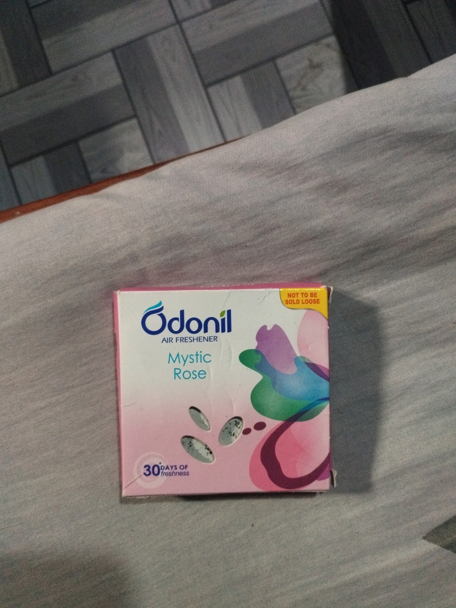 A square pink and white box of Odoril air freshener in Mystic Rose scent is centered in the frame. The box is placed on a light gray textured fabric. The background above the fabric shows a grey tiled floor arranged in a herringbone pattern. The air freshener box features the Odoril logo and "AIR FRESHENER" in blue text, and "Mystic Rose" in a lighter shade of blue. A yellow sticker in the upper right corner of the box reads "NOT TO BE SOLD LOOSE". The box also displays an illustration of colorful abstract flower petals and a circular icon with "30 DAYS OF freshness" written inside. There are no people or animals visible in the image. The scene is indoors. The location is not specified within the image.