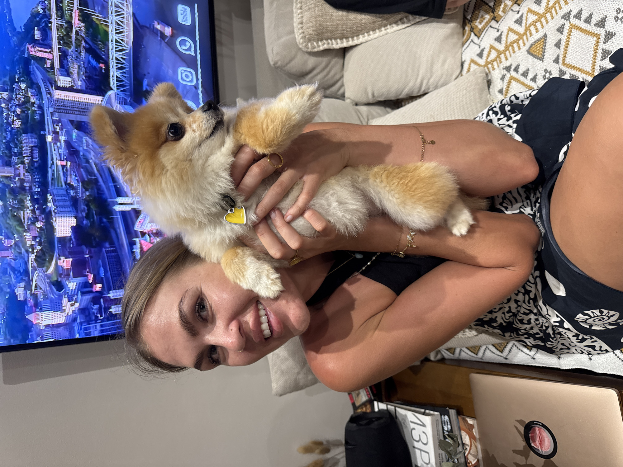 A smiling woman holds a tan and white Pomeranian dog. The dog wears a yellow heart-shaped tag on its collar. The woman wears gold rings and bracelets. Behind them is a television displaying an aerial cityscape. The location is Kuta Selatan, Indonesia. A laptop and books are on a surface next to them.