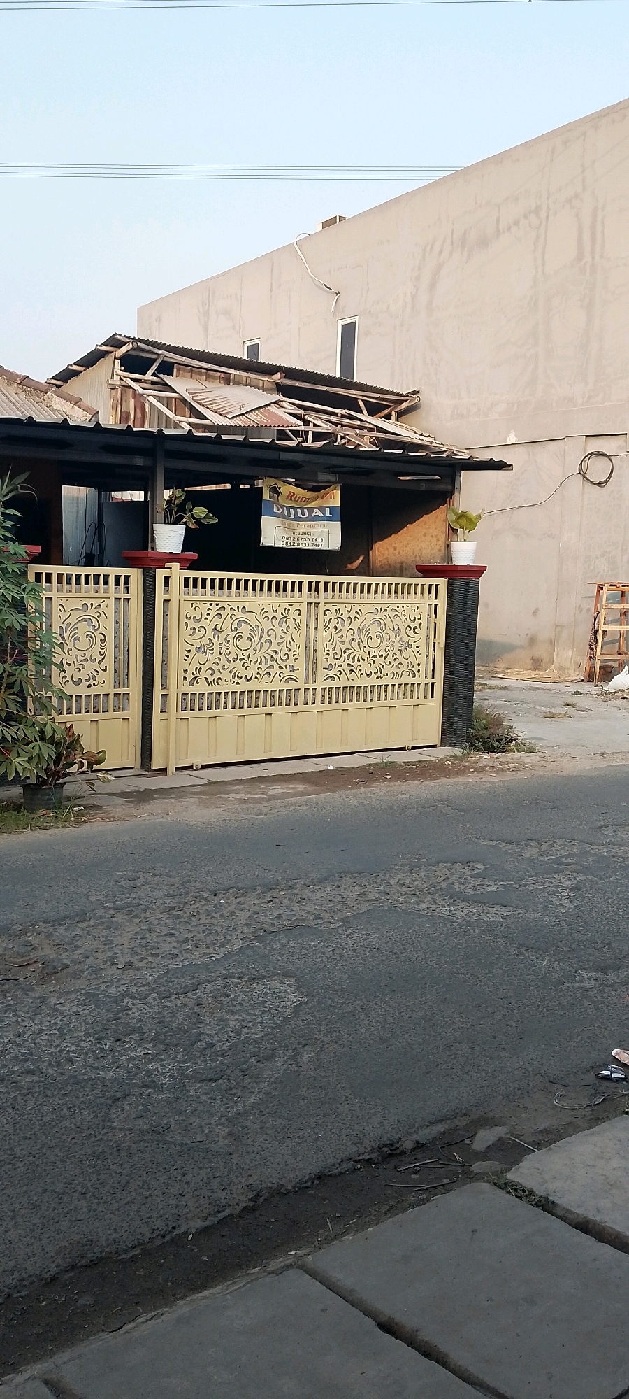In Satriajaya, Indonesia, a beige gate with ornate scrollwork stands before a building. A "Rumah Dijual" (House for Sale) sign hangs from the covered entrance of the building, which appears to be undergoing some disrepair with exposed roof framing and corrugated metal roofing. Two potted plants, one on each side of the entrance, add a touch of greenery. To the right, a tall, light-colored building with a rough, textured facade rises behind the smaller structure. A dislodged electrical conduit snakes across the wall of this larger building. In the foreground, an asphalt road shows signs of wear and tear, with patches of exposed aggregate. A concrete sidewalk runs along the edge of the road, and some debris is visible on its surface.