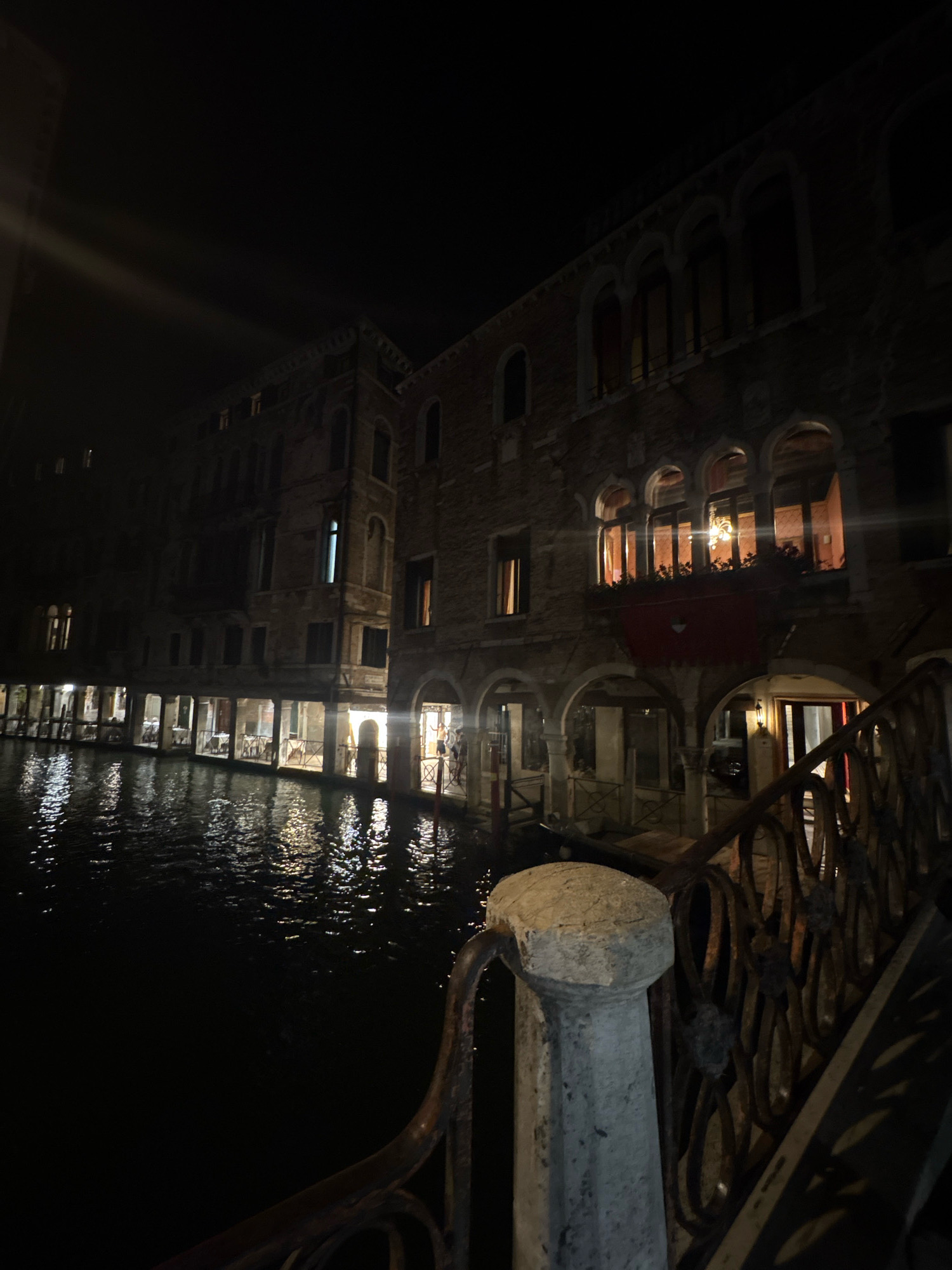 This is a nighttime exterior shot of a canal in Venice, Italy. The image captures a canal lined with historic buildings, their facades illuminated by interior lights. The water reflects the lights and the architecture, creating a shimmering effect. On the right side of the frame, the ornate metal railing of a bridge is visible in the foreground, with a stone post to its left. The buildings feature arched windows and balconies, with some windows glowing warmly from within. One building on the right has a red awning extending over an entrance. A person is visible standing near an entranceway on the left side of the frame. The scene is dark, with the primary illumination coming from the lights within the buildings and their reflections on the water.