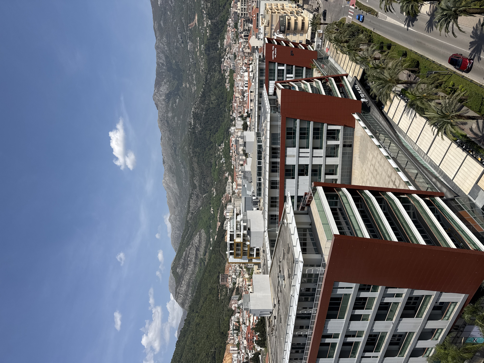 This high-angle panoramic photograph captures a vibrant coastal city, identified by context as Budva, Montenegro, set against a majestic mountainous backdrop under a clear blue sky. The image appears to be taken during the day, with bright sunshine casting distinct shadows from palm trees and buildings, indicating strong sunlight.
In the foreground, dominating the right side of the frame, are several modern, multi-story buildings featuring striking reddish-brown facade panels and numerous glass windows. One prominent building has "HOTEL TQ PLAZA" emblazoned vertically on its side, clearly indicating its purpose. Adjacent to this, a lower structure reveals the text "SHOPPING CENTER," suggesting a mixed-use complex. A wide, multi-lane road runs parallel to these buildings, adorned with tall palm trees that cast long shadows. Several vehicles, including a red SUV and a blue car, are visible on the road, hinting at active urban life.
Beyond these contemporary structures, the cityscape extends into the midground with a dense concentration of buildings, ranging from residential to commercial, many appearing to be older, terraced along the lower slopes of the mountains. The urban sprawl eventually gives way to a lush green forest, which then transitions into the rugged, grey mountains that form the imposing background, stretching across the entire width of the scene. The sky above is a brilliant azure, dotted with a few scattered white clouds. No individuals are visible in the busy urban and natural landscape. The overall scene depicts a dynamic interplay between modern development and breathtaking natural beauty.