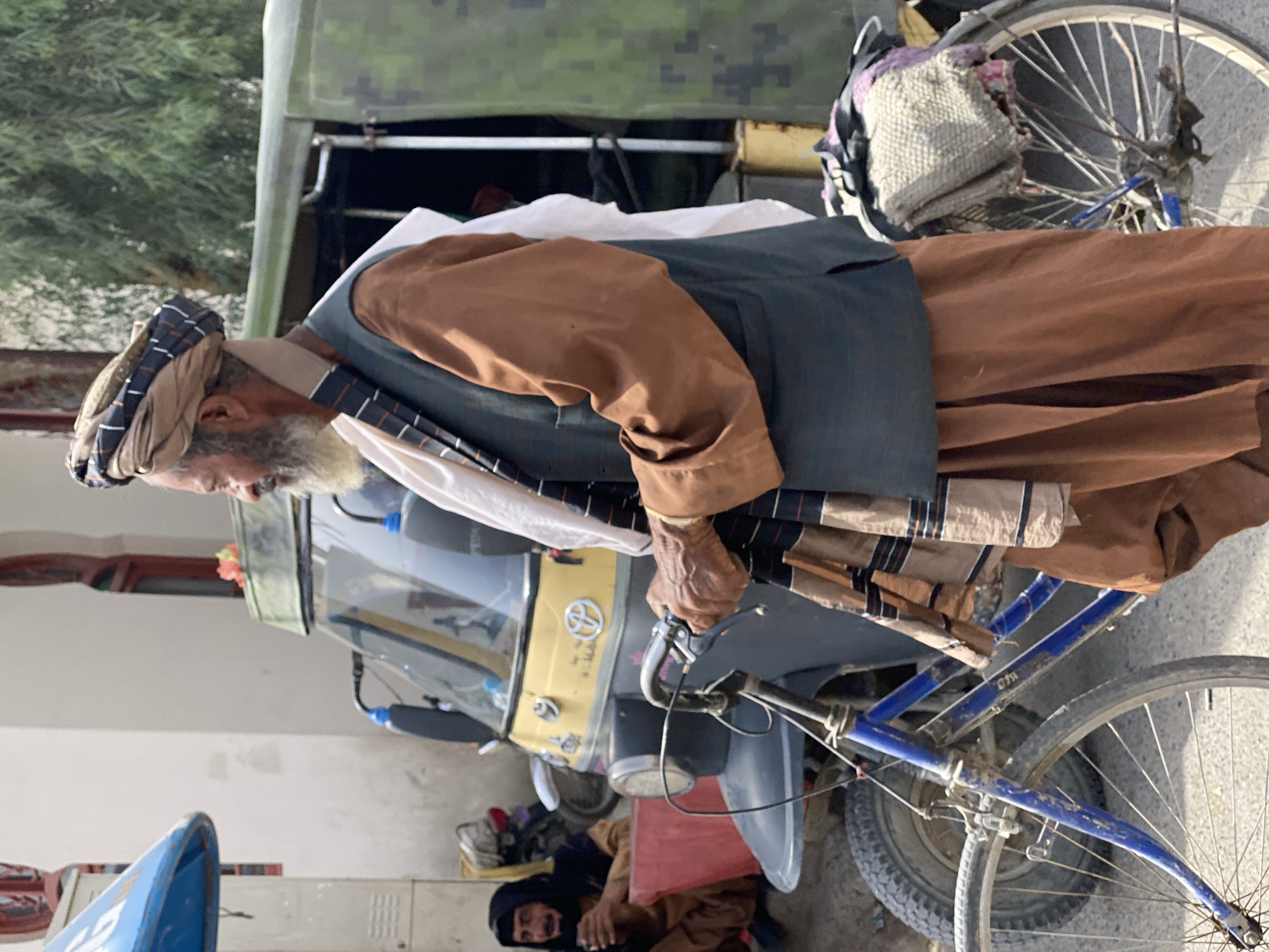 In the city of Kandahar, Afghanistan, an elderly man with a white beard and a turban stands by a bicycle and an auto-rickshaw. He is wearing a brown shirt, a dark vest, and loose brown pants. A striped scarf is draped around his neck. The auto-rickshaw, yellow and gray, is parked behind him, with a military-style green vehicle visible behind that. In the background, another man wearing a dark head covering and brown clothing is seated near a red bin, holding a cup. The environment appears to be an outdoor street scene with buildings and trees in the background.
