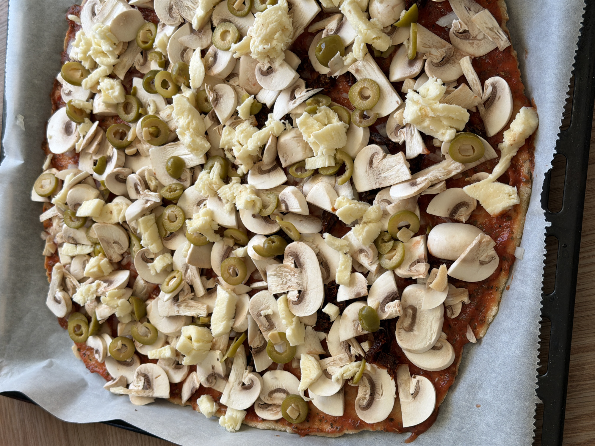 This close-up image displays an uncooked pizza, generously laden with toppings, prepared on a baking sheet lined with white parchment paper. The pizza base, which appears to be an irregular, possibly hand-formed crust, is evenly coated with a rich, reddish tomato sauce.
The surface of the pizza is densely covered with an array of ingredients. Numerous slices of fresh, white button mushrooms are distributed across the entire base, with many revealing their distinctive cap shapes and internal gill structures. Interspersed among the mushrooms are plentiful slices of green olives, adding a pop of color and an extra layer of flavor. A substantial amount of white cheese, which looks like roughly shredded or crumbled mozzarella, is scattered on top of the mushrooms and olives, with some pieces appearing larger and irregularly shaped.
The pizza occupies the central and dominant portion of the frame, indicating its readiness for baking. To the right, the black, corrugated edge of a baking tray is visible, while a light-colored wooden surface can be seen along the left and bottom edges of the image, beneath the parchment paper. The scene captures a moment of culinary preparation, likely in a home kitchen in Riga, Latvia, although no specific visual cues within the image itself confirm the city. There are no people or discernible text visible in the media.