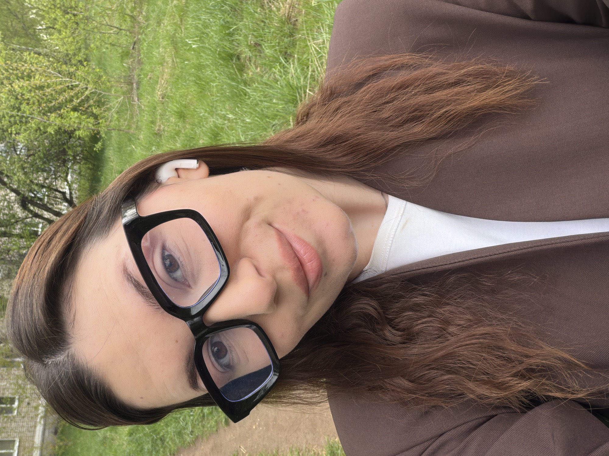 A selfie depicts a young woman in Chaikovsky, Russia. She is wearing a brown jacket, a white top, and black-framed glasses, and has an earbud in her ear. Her brown hair is styled loosely. The background shows greenery, suggesting a park or outdoor area within the city.