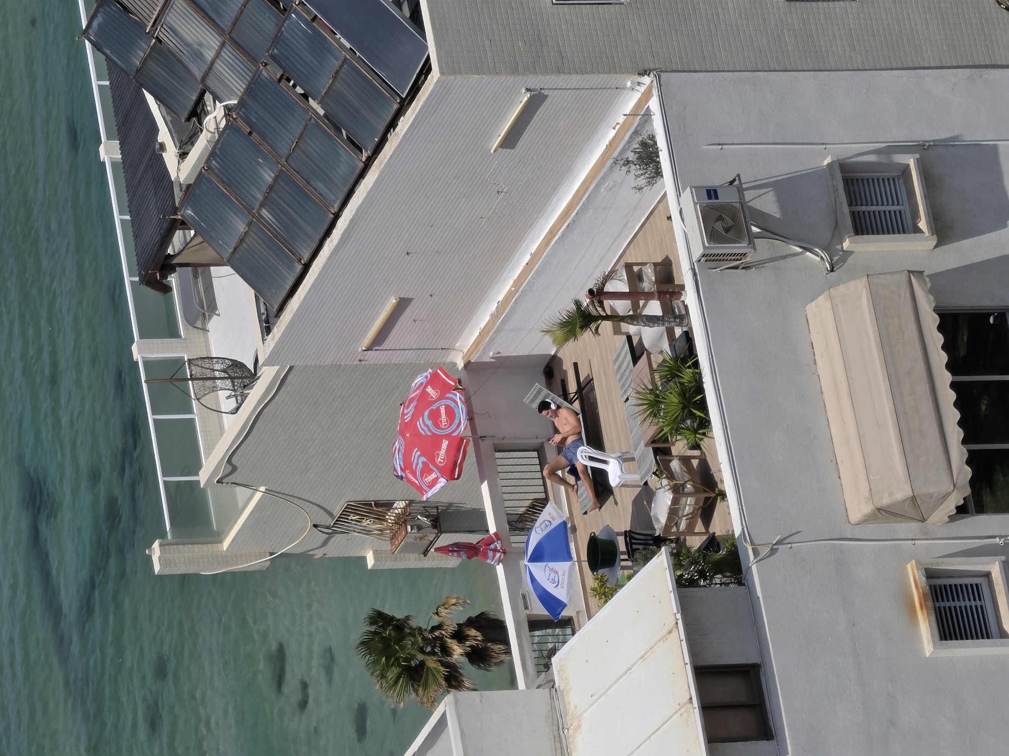 The image, taken from above, shows a rooftop terrace in Bat Yam, Israel. A man wearing headphones and blue shorts sits on a chair, looking at a device in his hands. Red and blue umbrellas provide shade. The terrace is furnished with chairs, tables, and potted plants. Solar panels are installed on the roof next to the terrace. Below, clear turquoise water laps the building's edge.