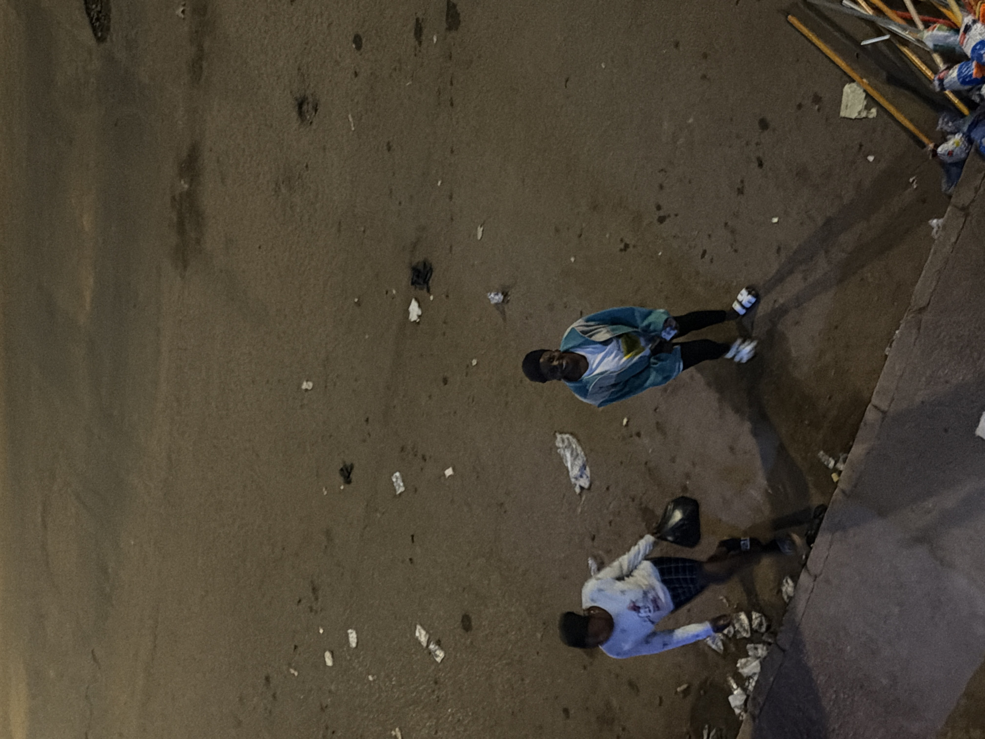 This image depicts an outdoor scene in the city of Bukuru, Nigeria. Two individuals are visible, standing on a dirt and pavement surface that is littered with debris, including paper and plastic waste.
The person on the left appears to be walking and is holding a dark plastic bag in their right hand. This individual is wearing a light-colored long-sleeved shirt with a graphic print, checkered shorts, and dark footwear.
The person on the right is standing and facing the camera. They are wearing a light-colored t-shirt underneath a teal-colored hooded jacket, black pants, and white slides or sandals.
The environment suggests an urban or semi-urban setting with evidence of untidiness. The ground is a mixture of dark, damp-looking soil or pavement, with lighter-colored debris scattered across it. In the lower right corner, a collection of objects, possibly including brooms and other tools, are leaning against a structure, and a trash bag is visible. The lighting indicates it may be evening or nighttime, with artificial light sources casting shadows.