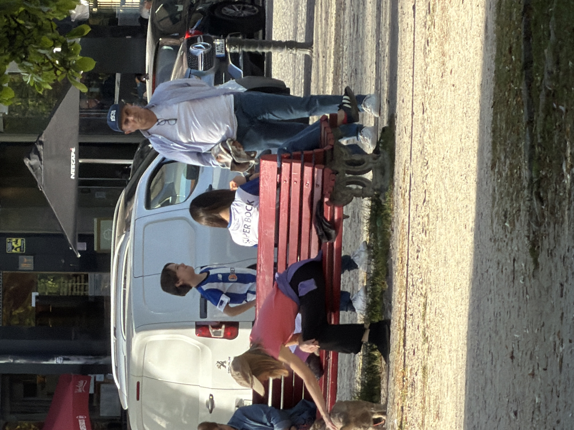 A group of people sits and interacts on a red park bench beside a paved path in a public space. A white van is parked behind the bench. A man in a white shirt, blue jeans, and a baseball cap sits on the bench with his legs stretched out across the bench. Two women are seated on the bench, with one leaning forward to pet a dog on the ground. A young man in a blue and white striped shirt is also seated on the bench. An open cafe/restaurant with a black awning and outdoor seating can be seen in the background, with a car parked near it. The paved path runs along the right side of the image, and a tree is in the upper left corner. Based on the license plates and architectural details, the location is consistent with the city of Porto, Portugal.