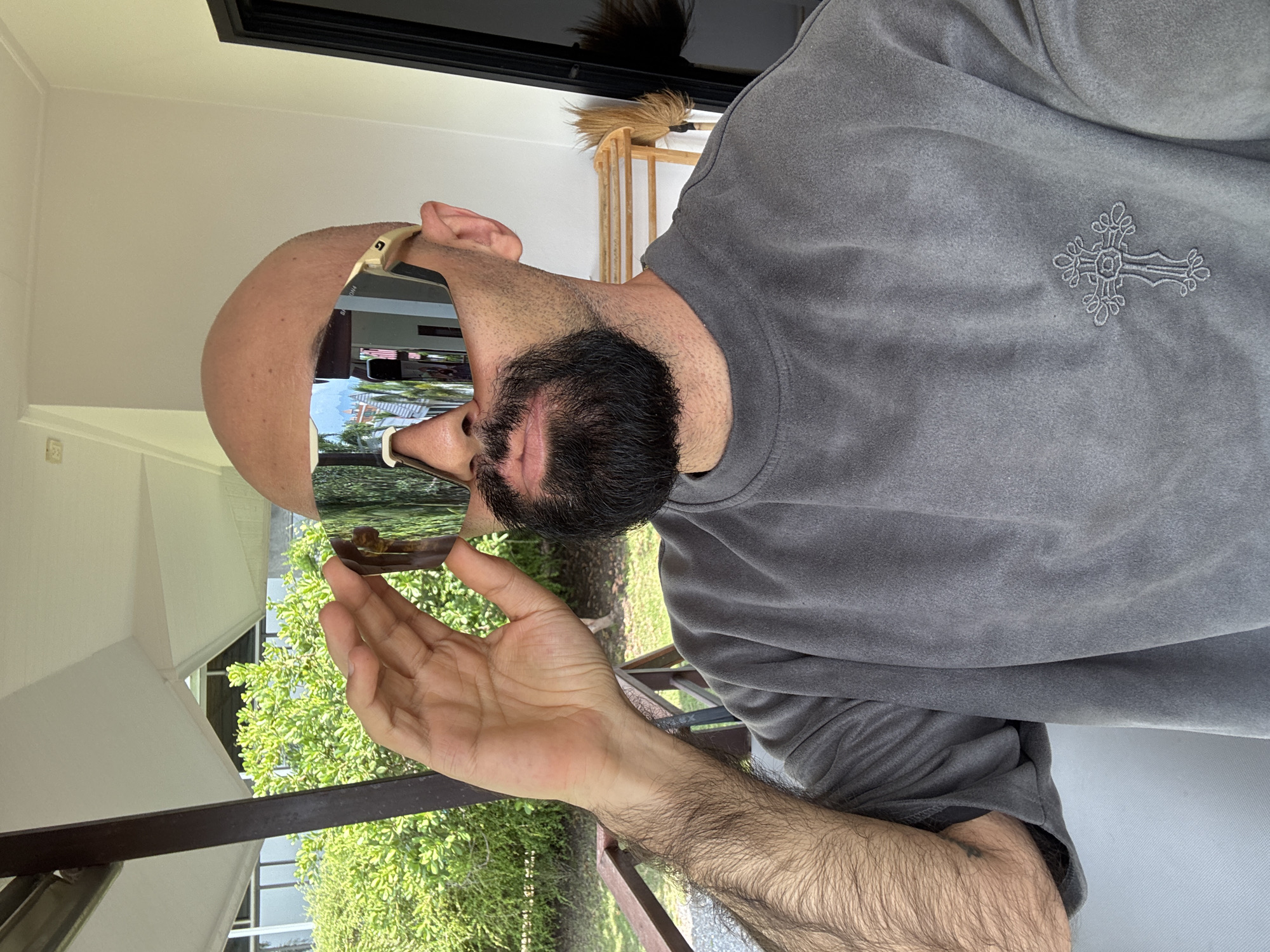 A bald man with a beard, wearing large mirrored sunglasses and a gray crewneck sweatshirt with a cross graphic, poses for a selfie on a balcony in a tropical outdoor setting. He is holding the frame of his sunglasses with his right hand. Lush green foliage and a portion of a building are visible in the background. The overall scene suggests a casual, sunny day in Ko Samui District, Thailand.