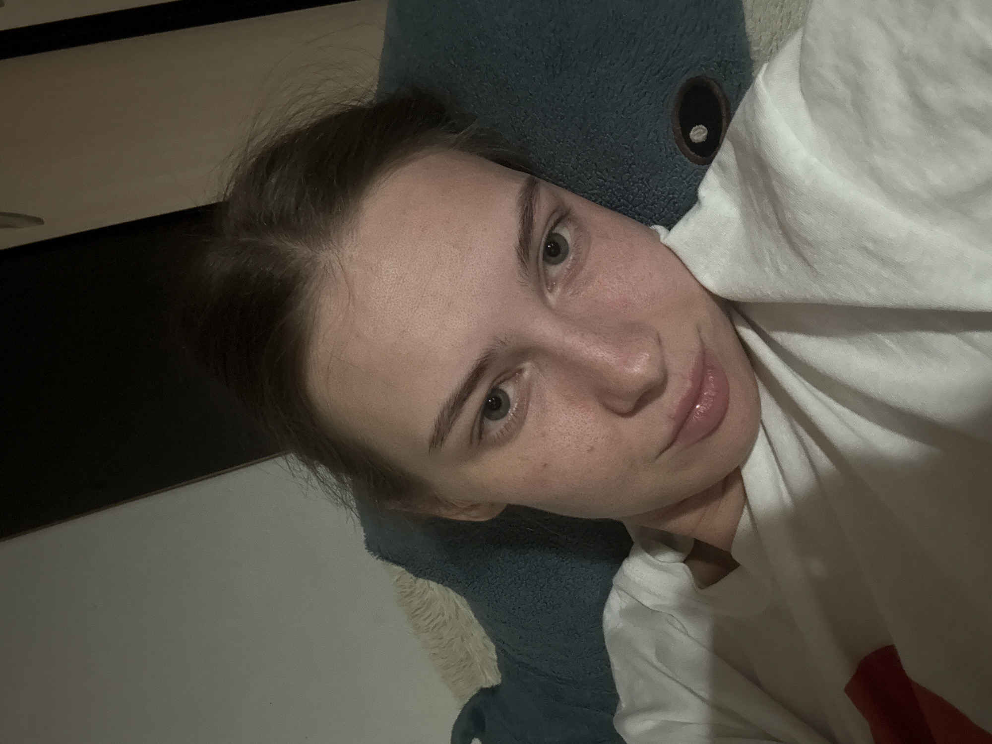 The image is a close-up, indoor selfie of a young Caucasian female with light skin, freckles, and light brown hair pulled back. She is lying down and looking at the camera. Her eyes are green, and her lips are slightly parted. She is wearing a white t-shirt. The background appears to be a bed with a blue pillow and a white blanket. The lighting is dim, suggesting it is nighttime or the room is dark. There is no discernible information about the location in the city of Perm, Russia, within the image.