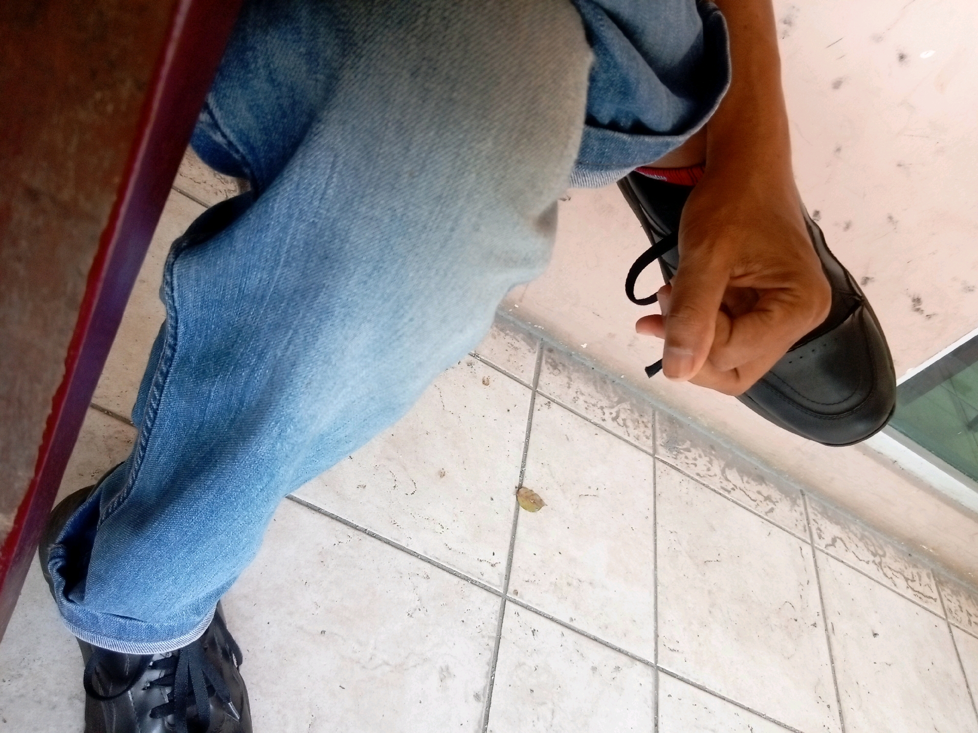 An overhead shot captures a person wearing light blue denim jeans and black dress shoes. The person's right hand is visible, adjusting the lace on the right shoe. The shoes are resting on a tiled floor. A wooden surface is partially visible on the left side of the frame. The scene appears to be an outdoor setting, possibly a patio or walkway, as indicated by the tiled floor and the presence of a fallen leaf. The lighting suggests it is daytime. There is no identifiable landmark or detail within the image that specifically places it in Batam, Indonesia.