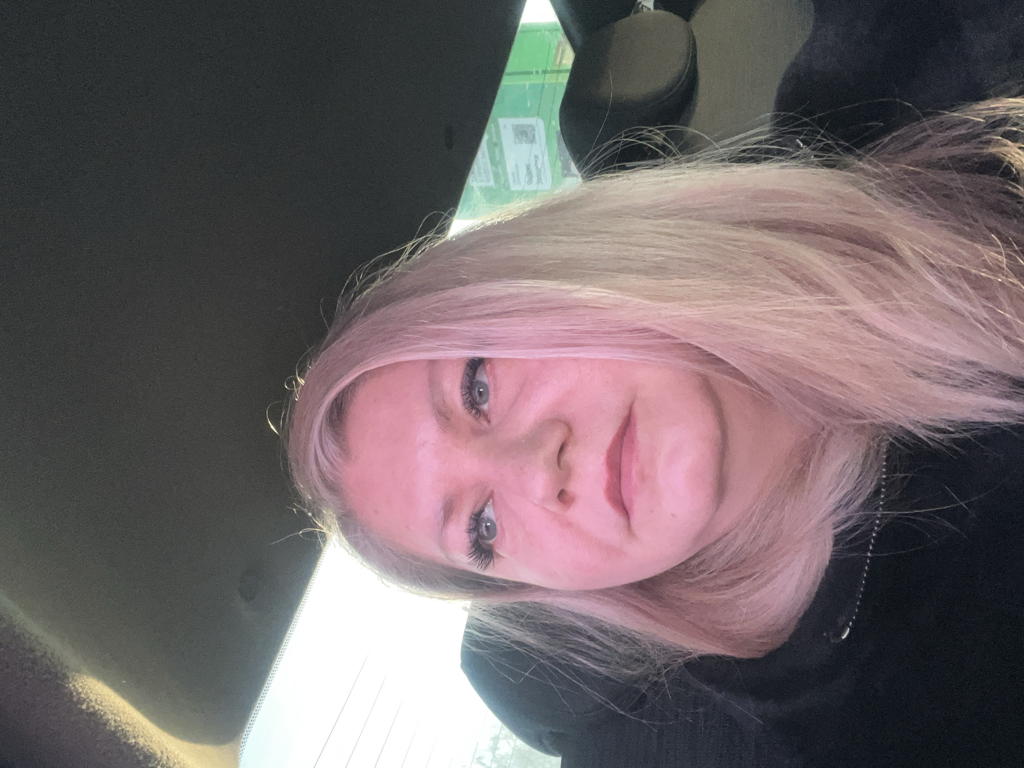 A female individual is depicted seated within a vehicle, observed in the city of Stary Chultem, Russia. She is positioned in what appears to be a passenger seat. Her long, light blonde hair exhibits a pinkish hue due to strong ambient light entering from the left. She is looking upwards and to her right. Her face shows defined eyelashes and lips with a reddish tint. She wears a dark, possibly black, zip-up garment with a visible metallic zipper pull. The vehicle's dark grey headliner is visible above, and a dark headrest is present behind and to her right. Through the vehicle's window, bright daylight is evident, along with faint horizontal lines resembling utility wires. In the background, a large green rectangular structure, possibly a building or sign, displays indistinct white graphical elements, including what may be a QR code.