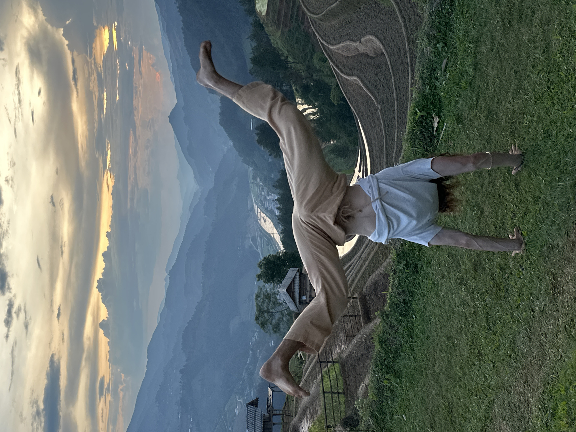 An individual is captured in an inverted, acrobatic pose on a patch of green grass, set against a majestic mountainous landscape in Mù Cang Chải, Vietnam. The person, an adult, is barefoot and wears loose light-colored pants and a pale t-shirt, which is slightly lifted. They appear to be mid-movement, perhaps in a handstand or a cartwheel, with their hands on the ground, one leg extended upwards, and the other bent at the knee.
Behind the person, distinct terraced rice fields form intricate patterns on the slopes. Beyond these agricultural terraces, a vast panorama of undulating mountain ranges stretches into the horizon, fading into soft blue and grey tones. A valley containing what appears to be a river or stream is visible between the distant mountains. In the middle ground, nestled among trees, a small, rustic wooden house adds a touch of human presence to the natural grandeur.
The scene is illuminated by the warm, soft light of either sunrise or sunset, with the sky showcasing a blend of golden, orange, and yellow hues mingling with dramatic blue-grey clouds. The overall impression is one of agility and freedom harmonized with the expansive, beautiful environment. No text is visible in the image.
