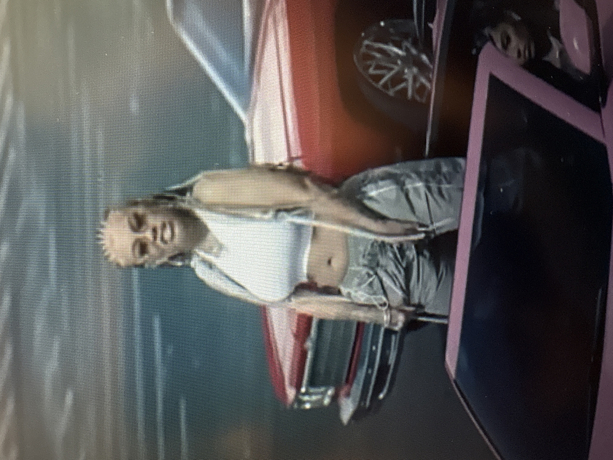 A medium shot captures a woman with blonde braided hair and hoop earrings, wearing a white crop top and gray athletic pants, standing next to a red vintage car. Her facial expression is neutral. In the foreground, a pink car's interior is visible, with another woman seated inside, looking to the left. The background appears to be an outdoor setting, possibly a street, with a sky that suggests overcast conditions. The image quality is low resolution, suggesting it may be a screenshot or a photograph of a screen. There are no discernible landmarks or features that would indicate the location as Sofia, Bulgaria.