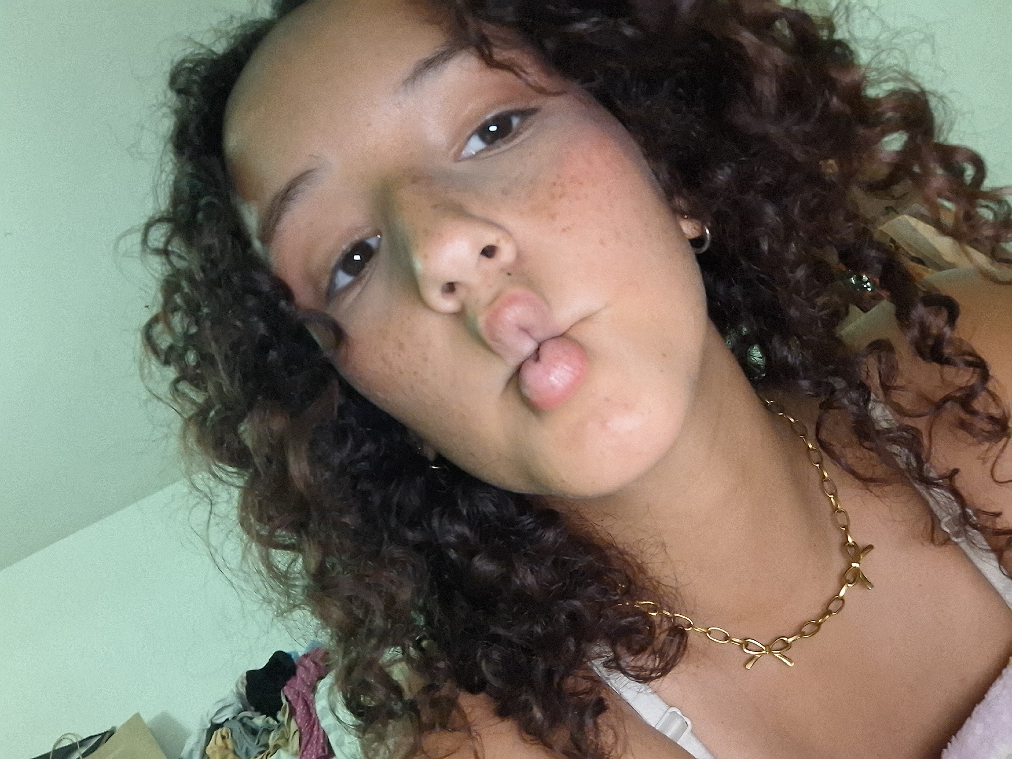 A close-up, overhead shot captures a young person with curly, dark brown hair pursing their lips for a selfie. Their skin is adorned with freckles, and they are wearing a delicate gold chain necklace with bow charms. A white strap from an undergarment is visible on the left side of the frame. The background is a light green wall, with a pile of clothes and a bag on the far left. The lighting is soft, creating a casual and intimate atmosphere. The image does not contain any information that allows for the determination of its location within Juan Griego, Venezuela.