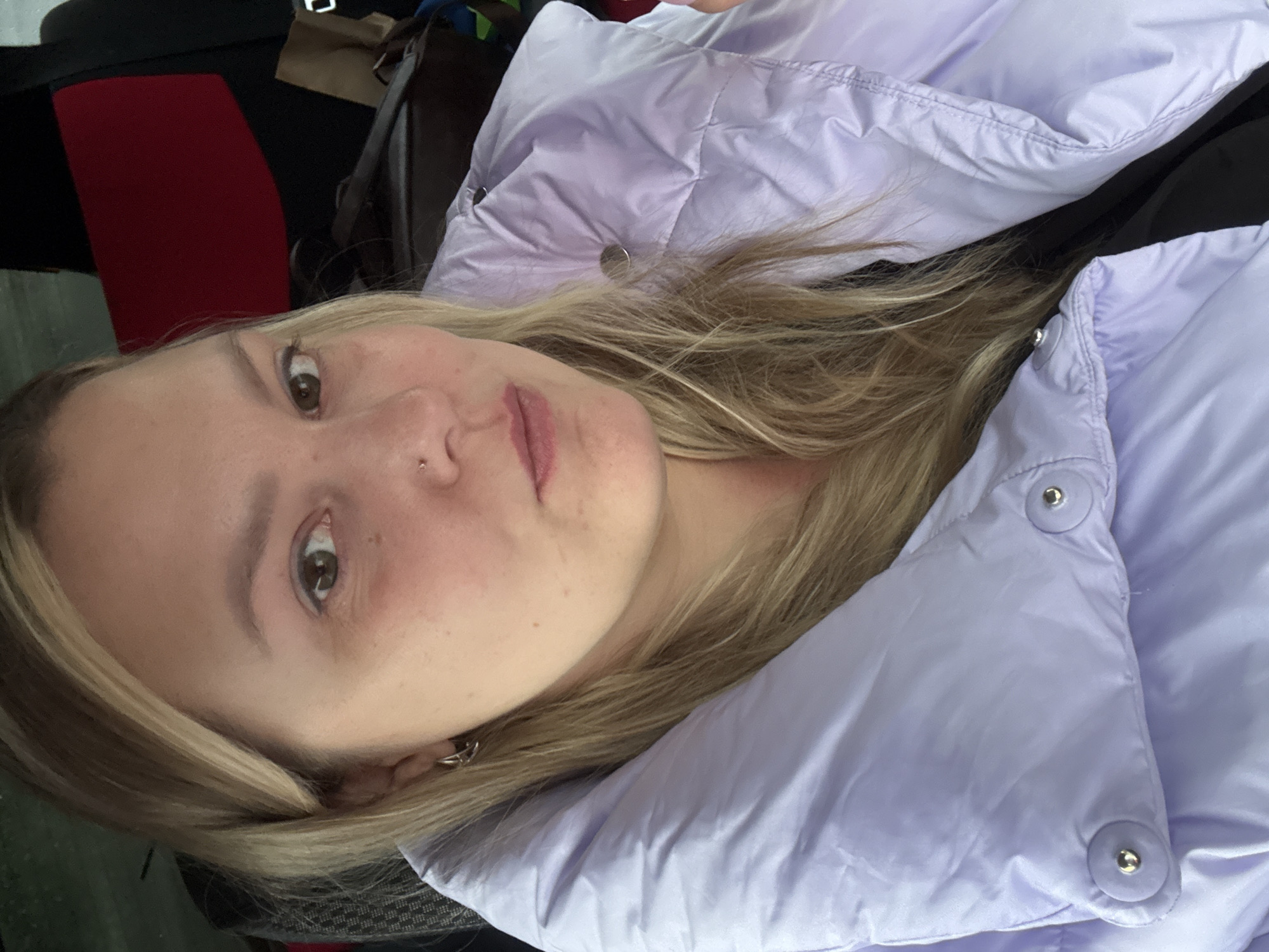 A woman is photographed indoors, wearing a light purple puffer jacket. Her hair is blonde. The image is slightly rotated counter-clockwise. The background shows part of a red and black seat, and what appears to be a bag. The location cannot be determined to be Chaikovsky, Russia, based solely on this image.