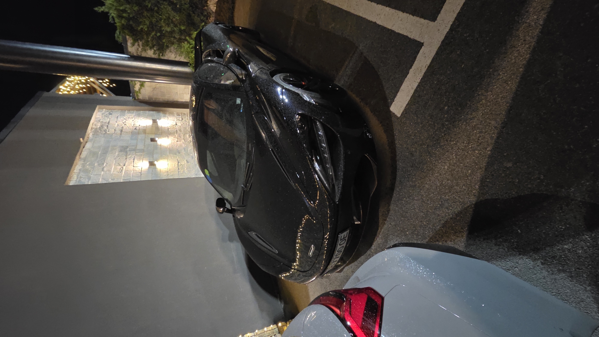 Here is a forensic description of the image:
The image depicts two vehicles parked at night in Unknown, Unknown. The dominant vehicle is a black McLaren, situated at an angle to the frame. Raindrops are visible on its surface, indicating recent or ongoing precipitation. Partially visible in the foreground is another car, light gray in color, with a red tail light illuminated. The vehicles are parked in a designated parking spot marked with white lines on the asphalt. To the left of the McLaren is a gray wall with embedded lights and stone cladding on a portion of the wall. An overhead lighting fixture is partially visible above the wall. Green foliage is also present in the background, near the light fixture. The overall lighting of the scene is dim, with light sources primarily from the building and the headlights of unseen vehicles.