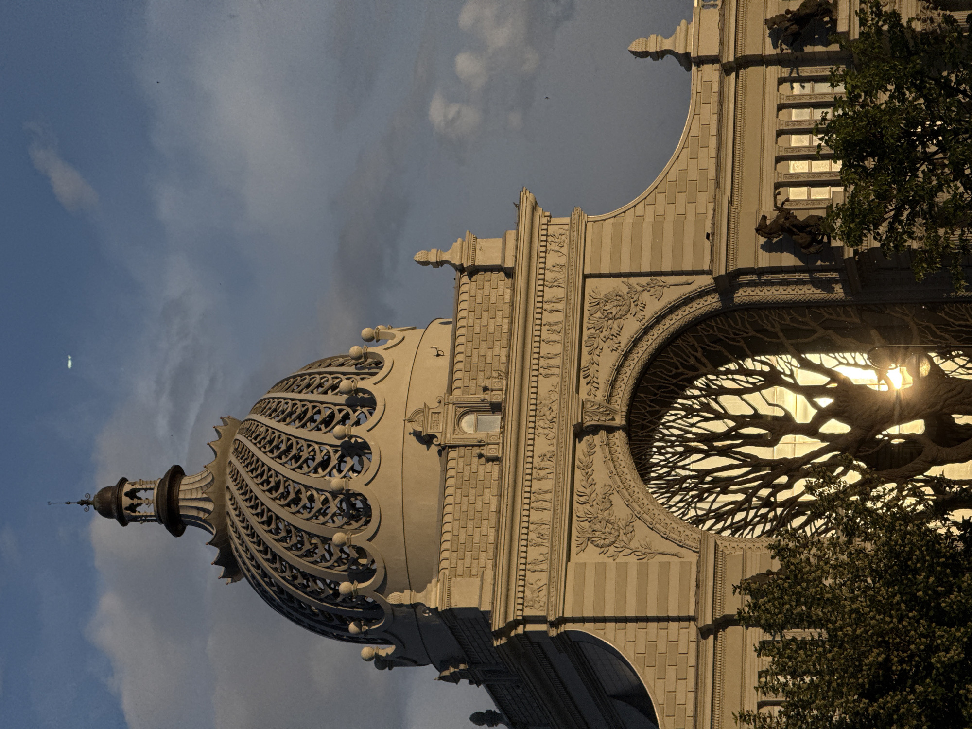 The image shows the Palace of Farmers in Kazan, Russia, tilted for an unconventional perspective. The building's dome, an ornate openwork structure, occupies the upper left. Adjacent is the façade, with brick-like textures, decorative carvings, and statues. A prominent feature is a large, arched window containing a sculpted tree with branches. Sunlight glints through the window and the branches, partially obscured by green foliage from an actual tree. The background reveals a sky with clouds.