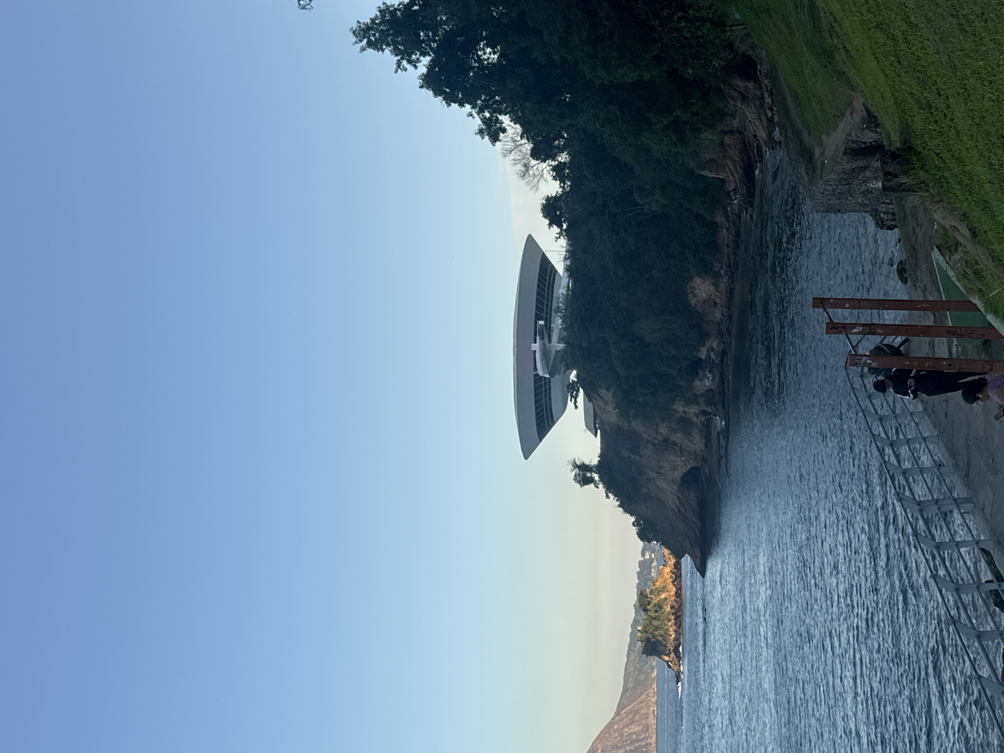 The image shows the Museum of Contemporary Art (MAC) in Niterói, Brazil, designed by Oscar Niemeyer. The museum is a white, saucer-shaped building perched atop a cliff overlooking the Guanabara Bay. The scene is captured during daylight, with clear blue skies and calm water. In the foreground, a walkway lined with a metal railing follows the coastline. Two individuals, one standing and one seated, are visible near a set of outdoor exercise equipment on the walkway. The cliff face is covered with dense green vegetation, and in the distance, a mountainous landscape can be seen across the bay. The overall atmosphere is serene and picturesque, highlighting the architectural marvel against the natural beauty of the coastal landscape.