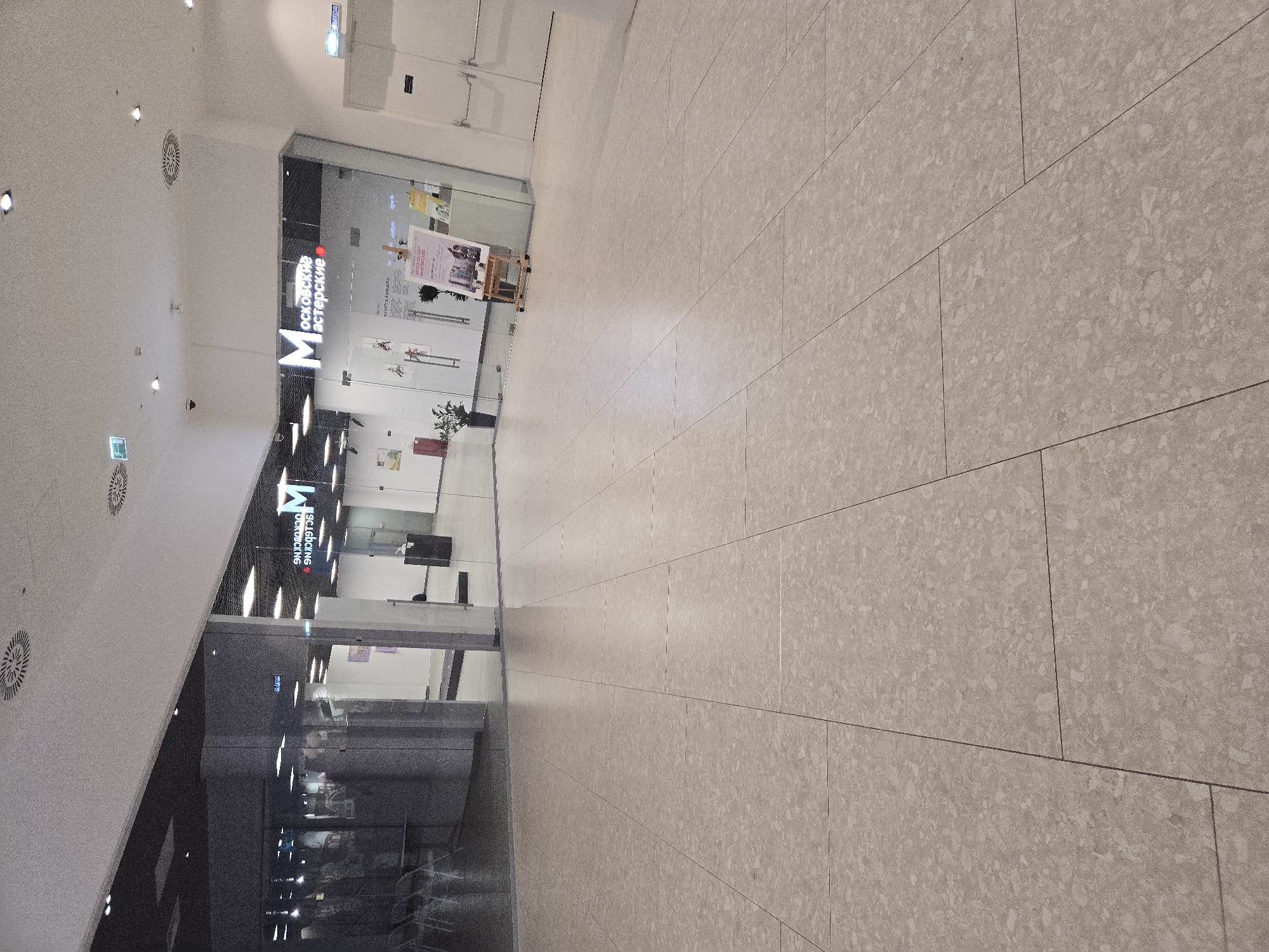 The image captures an interior view of a modern commercial space, likely a shopping mall or a public building, with a focus on the floor and the entrance to a business. The floor is tiled with large, light-colored, speckled tiles laid in a grid pattern. In the background, through a glass facade, is the entrance to a store or workshop with signage that reads "МОСКОВСКИЕ МАСТЕРСКИЕ" (Moskovskie Masterskie), indicating it is located in Moscow, Russia. The signage features a large white "M" followed by the text in red. Inside the establishment, a display easel with a poster is visible, along with some artwork or exhibits. To the left of the entrance, there's a dark grey column and a mirrored wall reflecting the interior. The environment appears clean and well-lit, with ceiling lights and ventilation grilles visible. The scene is devoid of any people actively engaged in the frame.