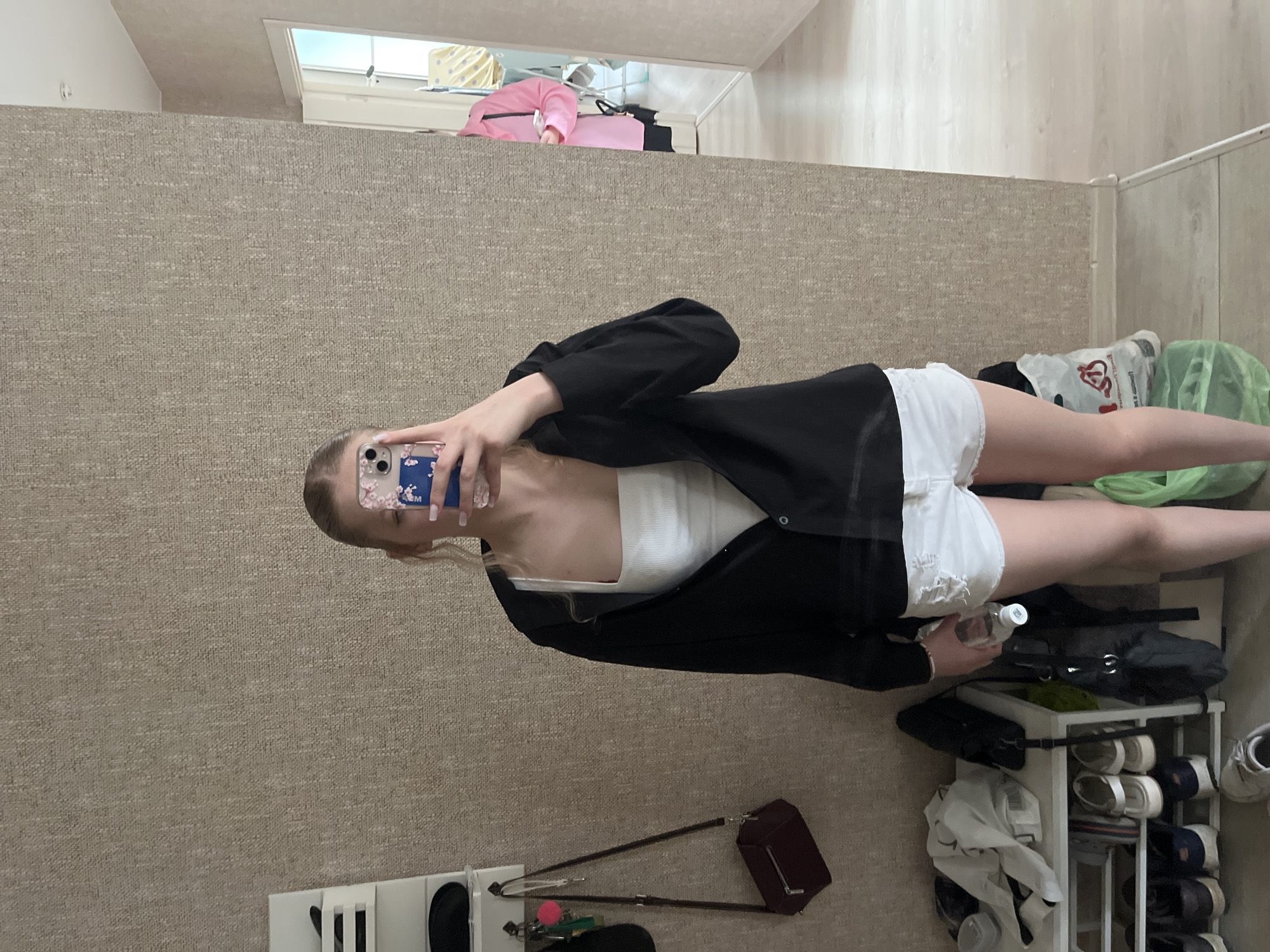The image shows a young Caucasian woman with blonde hair pulled back in a ponytail, taking a mirror selfie. She is wearing a white tank top, an open black button-down shirt, and distressed white denim shorts. She is holding a clear water bottle in her right hand. Her left hand is holding a smartphone with a floral case.
In the background, to the left, is a white shelving unit with various items, including bags, shoes, and what appears to be a dark red handbag hanging from a hook. To the right of the woman, a doorway leads to another room where another person, wearing a pink shirt and black shorts, is visible. The setting appears to be an indoor space, possibly an apartment or house, with textured wallpaper and a wooden floor. The presence of a shoe rack and items like bags suggests a residential area. The image has a neutral lighting condition. No specific elements in the image conclusively place the scene in Perm, Russia.