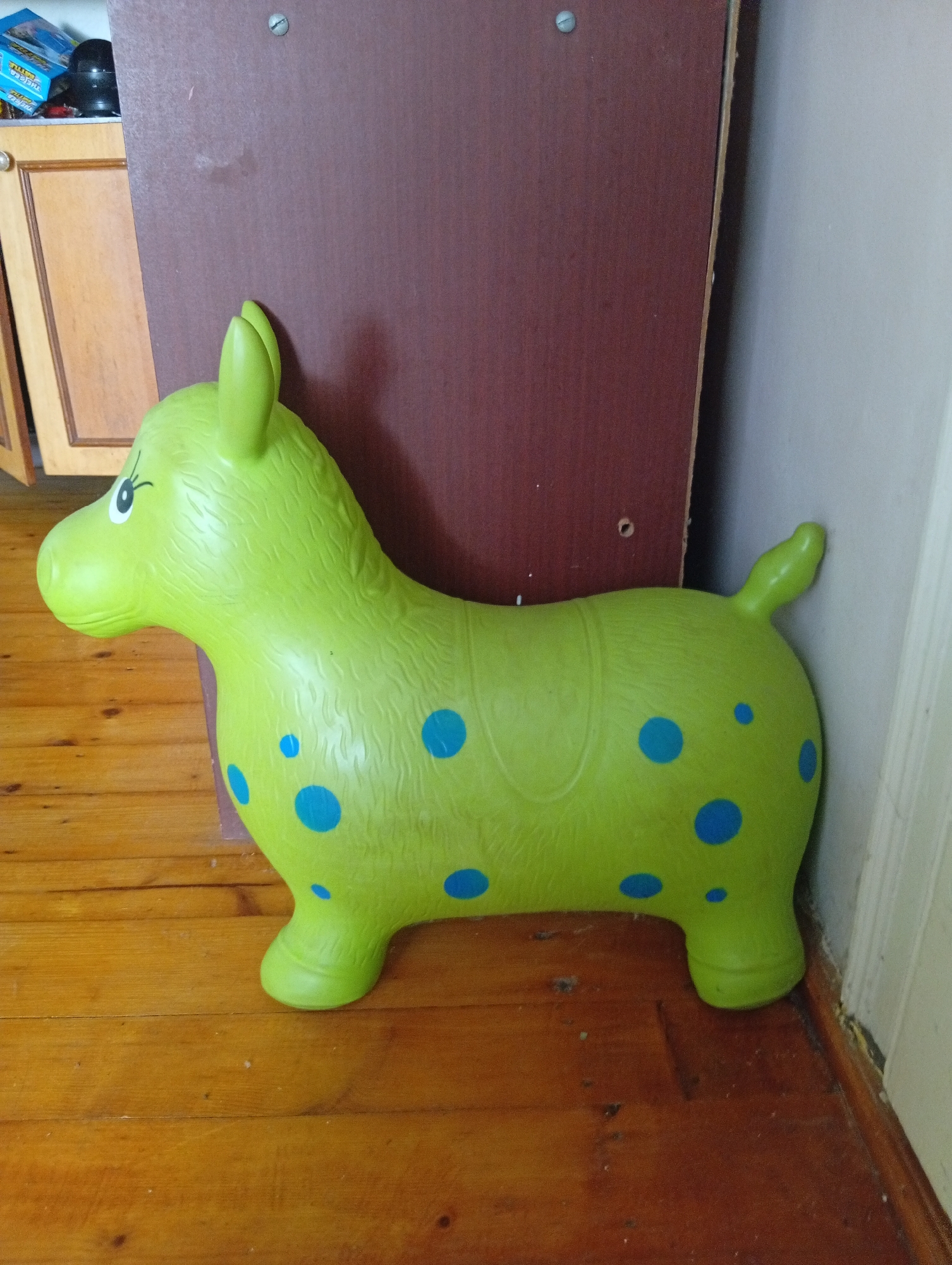 A bright green, inflatable horse toy with blue polka dots sits on a wooden floor. The toy is positioned in the foreground, facing left. Behind the toy, a dark red, wooden cabinet is visible, with one of its doors slightly ajar. To the right of the cabinet is a white wall. The floor appears to be made of polished wooden planks. The overall scene suggests an indoor setting, possibly a child's play area or a room in a house. The image does not provide any specific clues to its location in Veliatyno, Ukraine, other than it being a static indoor environment.