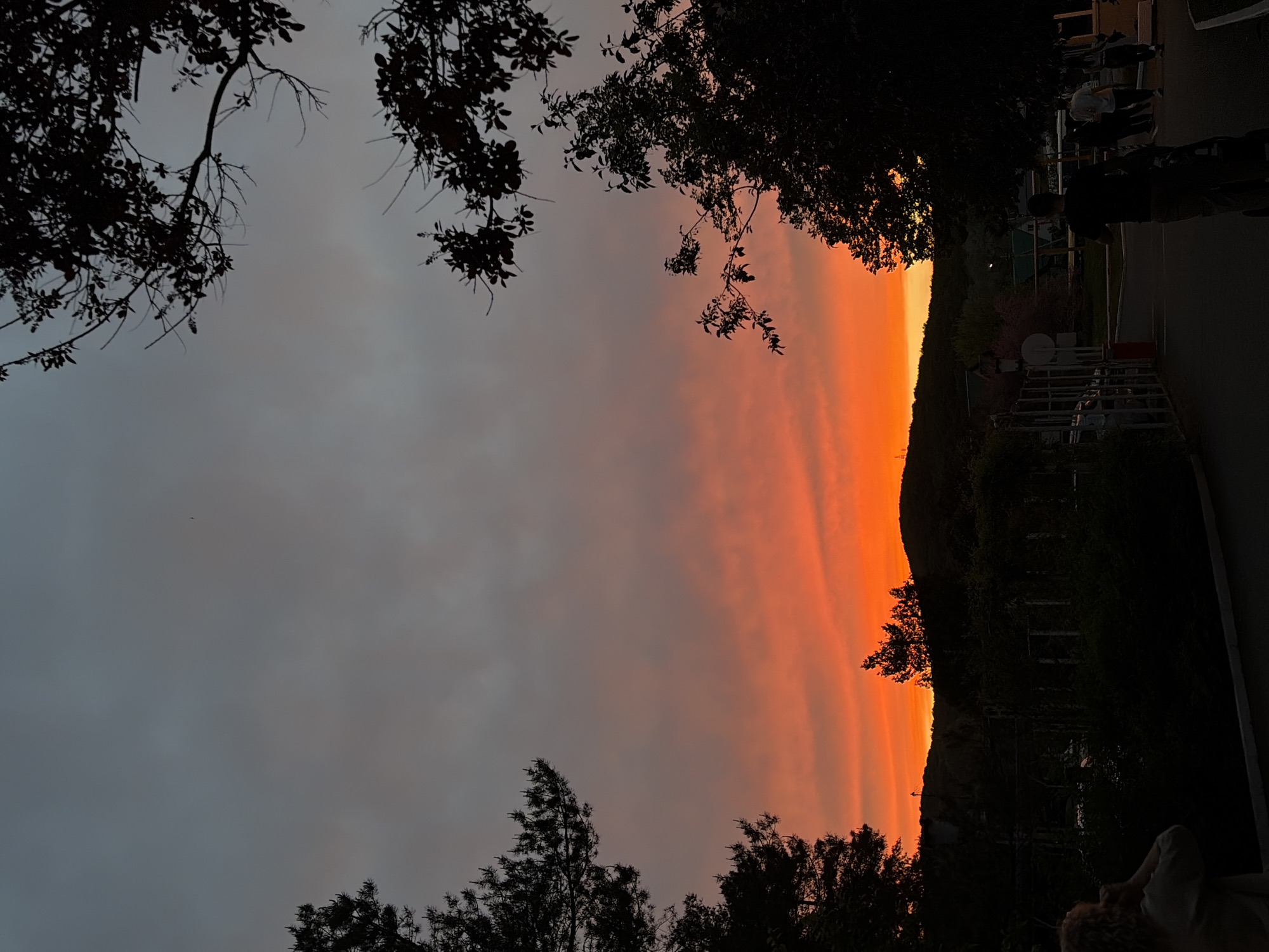 The image shows a sunset in Goryachiy Klyuch, Russia. The sky transitions from gray on the left to vibrant orange on the right, indicating the setting sun. Dark silhouetted trees frame the top and bottom of the image, with branches extending into the sky. On the right side of the image, a dark hill or mountain is visible. People are present on the right side of the image. A metal railing and possibly a road can be seen.