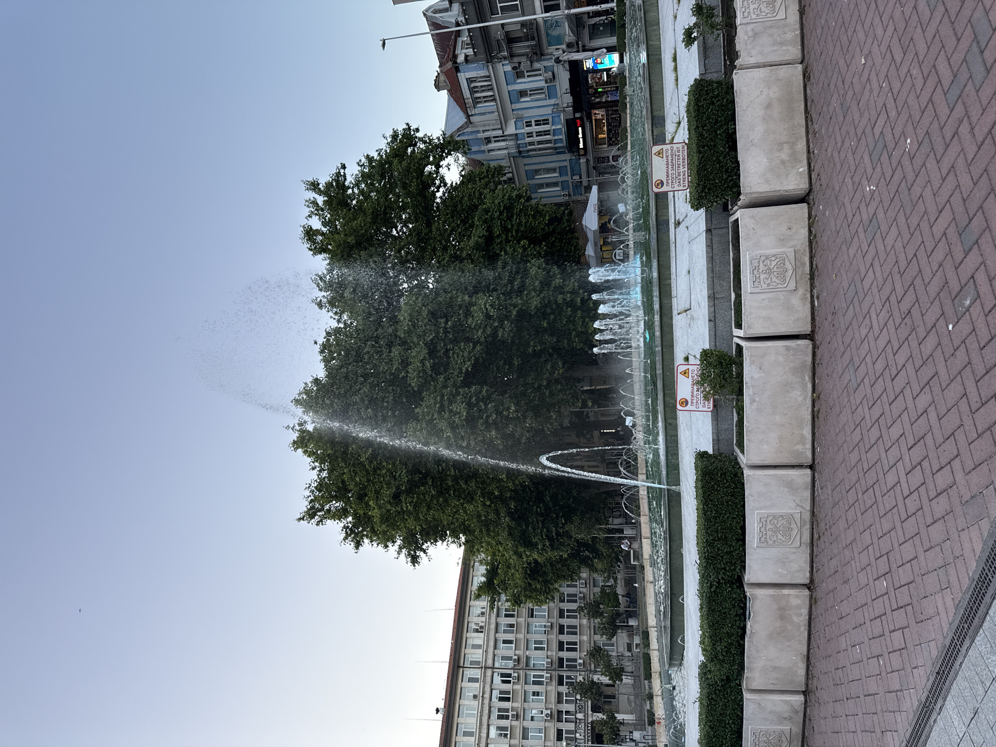 A wide shot shows a fountain in an urban square, likely in the city of Varna, Bulgaria, given the emblem on the planters and signs in Bulgarian. The fountain, with multiple jets of water illuminated by blue light, is centrally located in a circular basin surrounded by a low wall. Beyond the fountain, a large building with many windows and air conditioning units dominates the background, with other buildings to its right. Trees line the edge of the fountain and surround it. In the foreground, there are large concrete planters with manicured bushes and decorative emblems. The ground is paved with red bricks. Several signs are visible near the fountain, some with warnings in Bulgarian. The sky is clear and light blue, suggesting it is either early morning or late afternoon. There are no people clearly visible in the foreground, but a few figures can be seen in the background, sitting or walking. A small dark object, possibly a drone, is visible in the sky in the upper left quadrant.