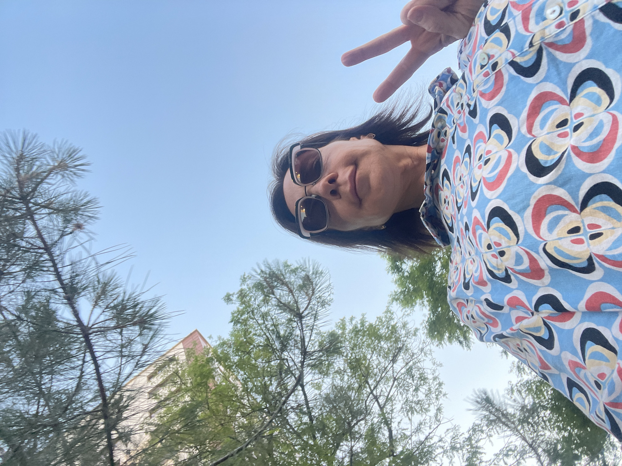 A low-angle outdoor selfie captures an individual in Sochi, Russia. The central figure is a middle-aged woman with dark hair, wearing large-framed sunglasses featuring a light-colored upper rim. She is clad in a light blue button-up shirt patterned with abstract floral or geometric designs in red, black, off-white, and blue. Her left hand is raised, forming a peace sign with the index and middle fingers. The background consists of a clear, light blue sky. To the left, green foliage from both needle-leaf (possibly pine) and broadleaf trees is visible, partially obscuring a light-colored building with visible windows and a sloped roofline in the upper left quadrant.